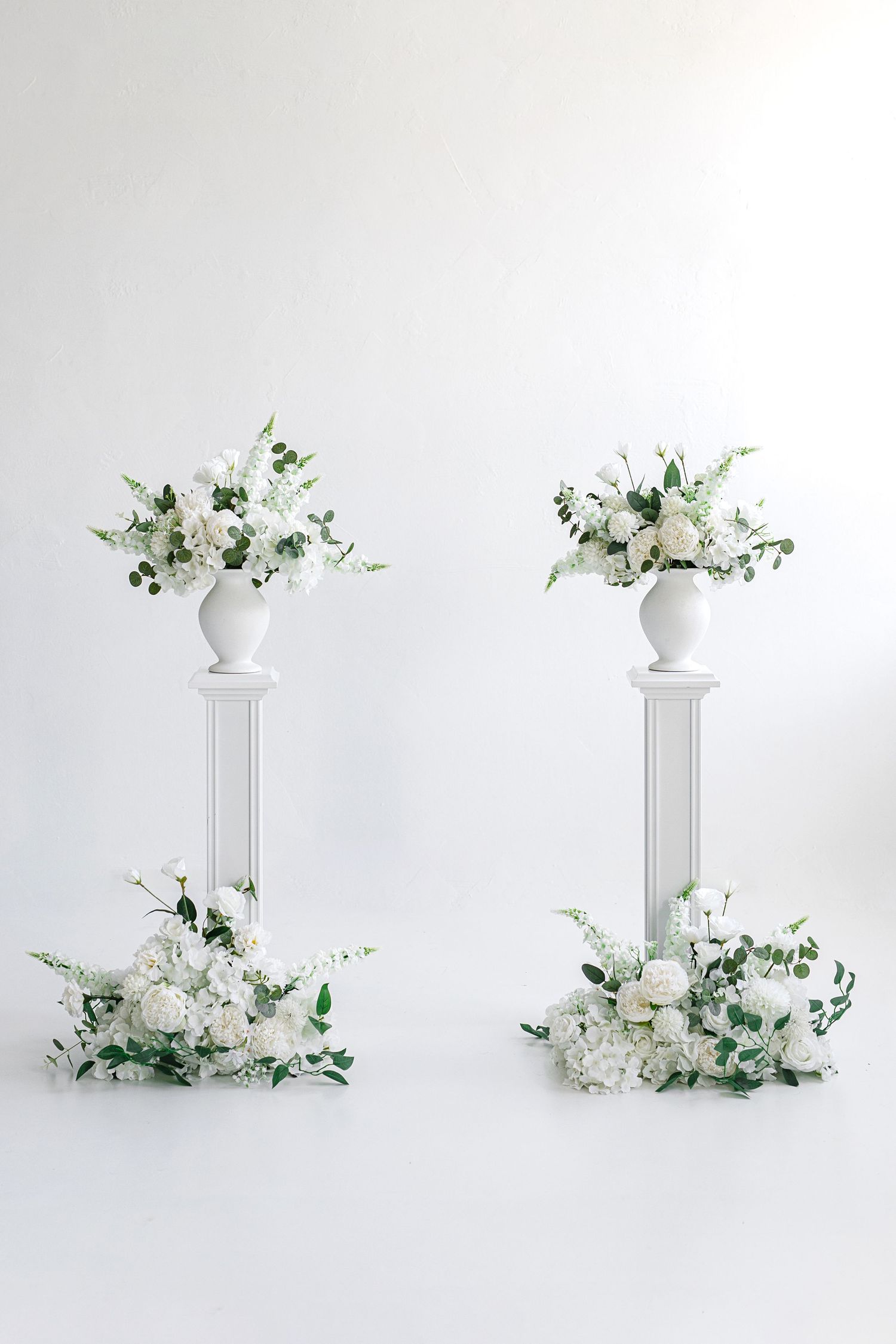 Two white pedestal columns adorned with elegant white floral arrangements and greenery against a white background.