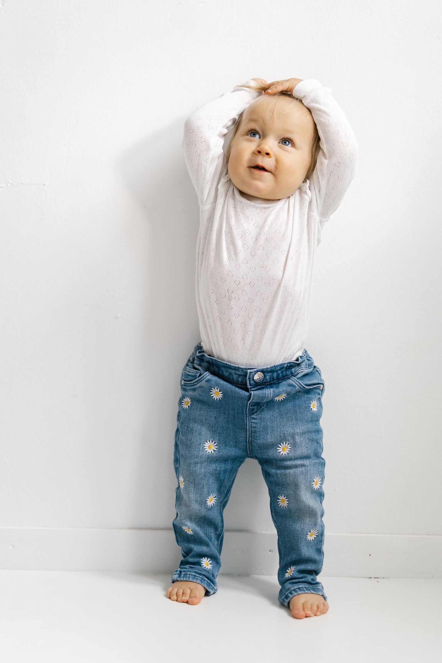 Adorable baby standing against white wall, wearing white bodysuit and daisy-print jeans, hands raised above head.