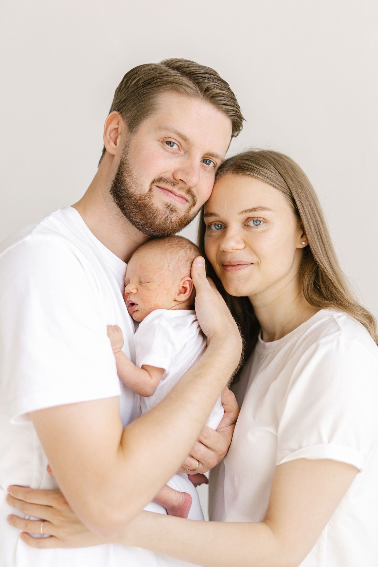 Happy young parents in white t-shirts holding their sleeping newborn baby, posing for a family portrait.