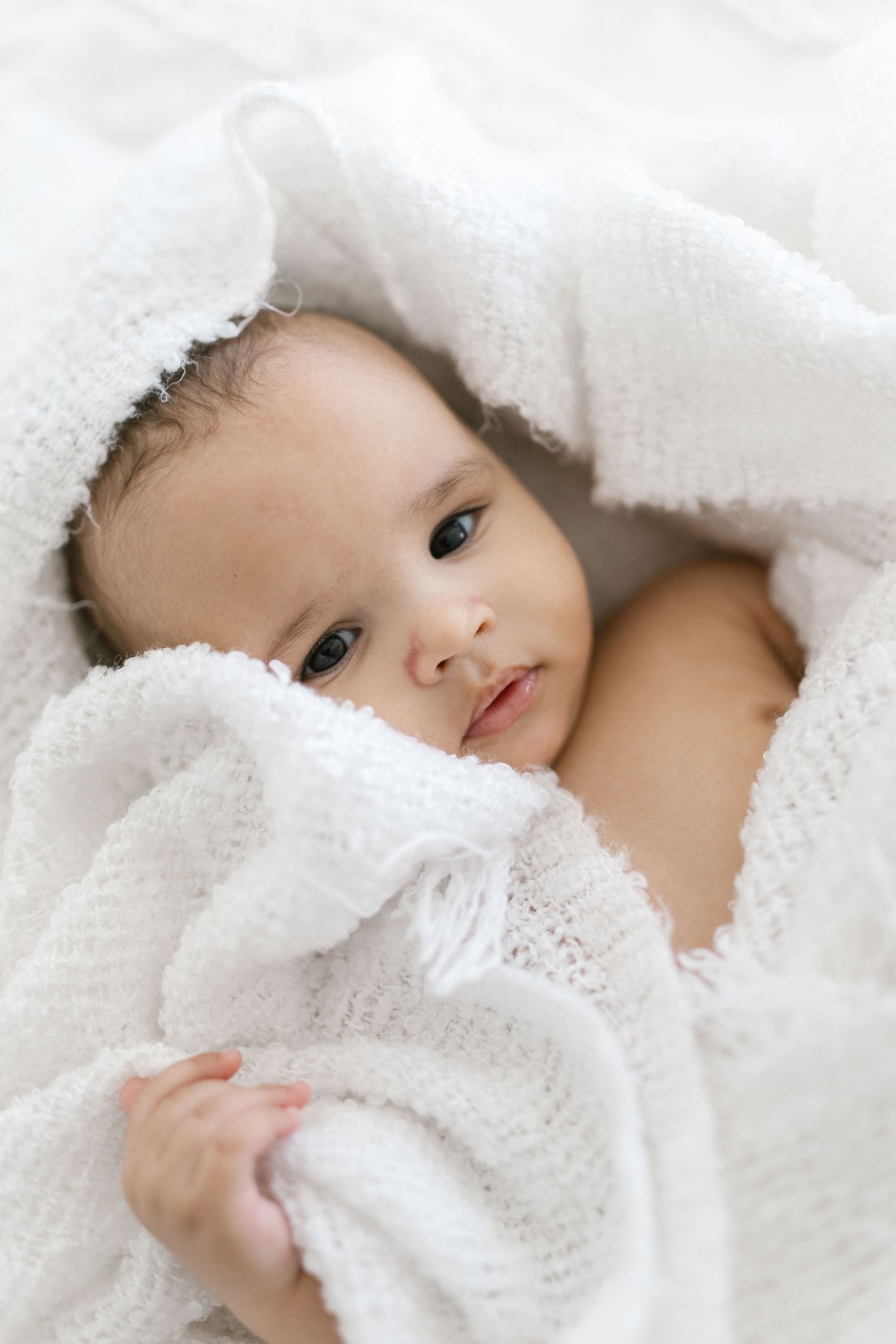 A cute baby wrapped snugly in a soft white knit blanket, gazing curiously with bright eyes.