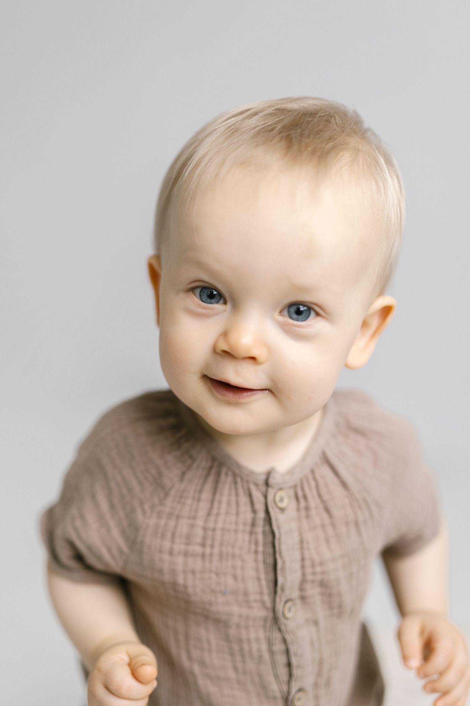 Baby in beige outfit smiling against light gray background.