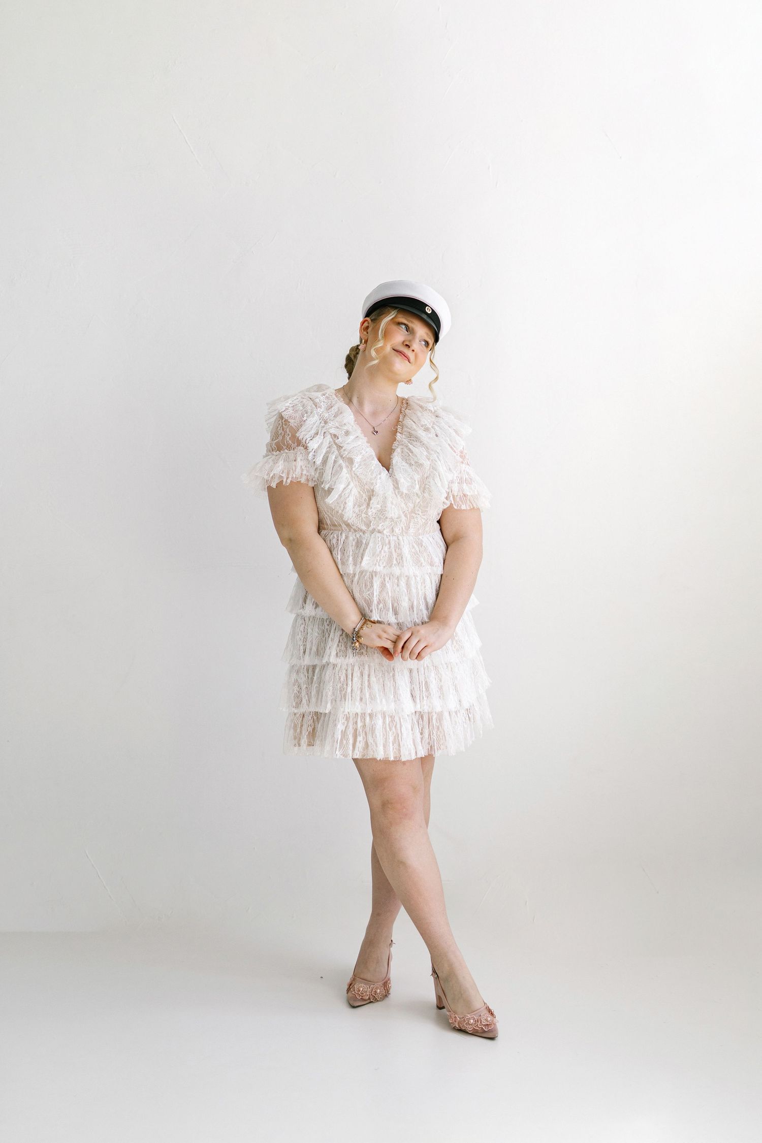 Woman in white lace ruffle mini dress and white cap poses gracefully against a white background.
