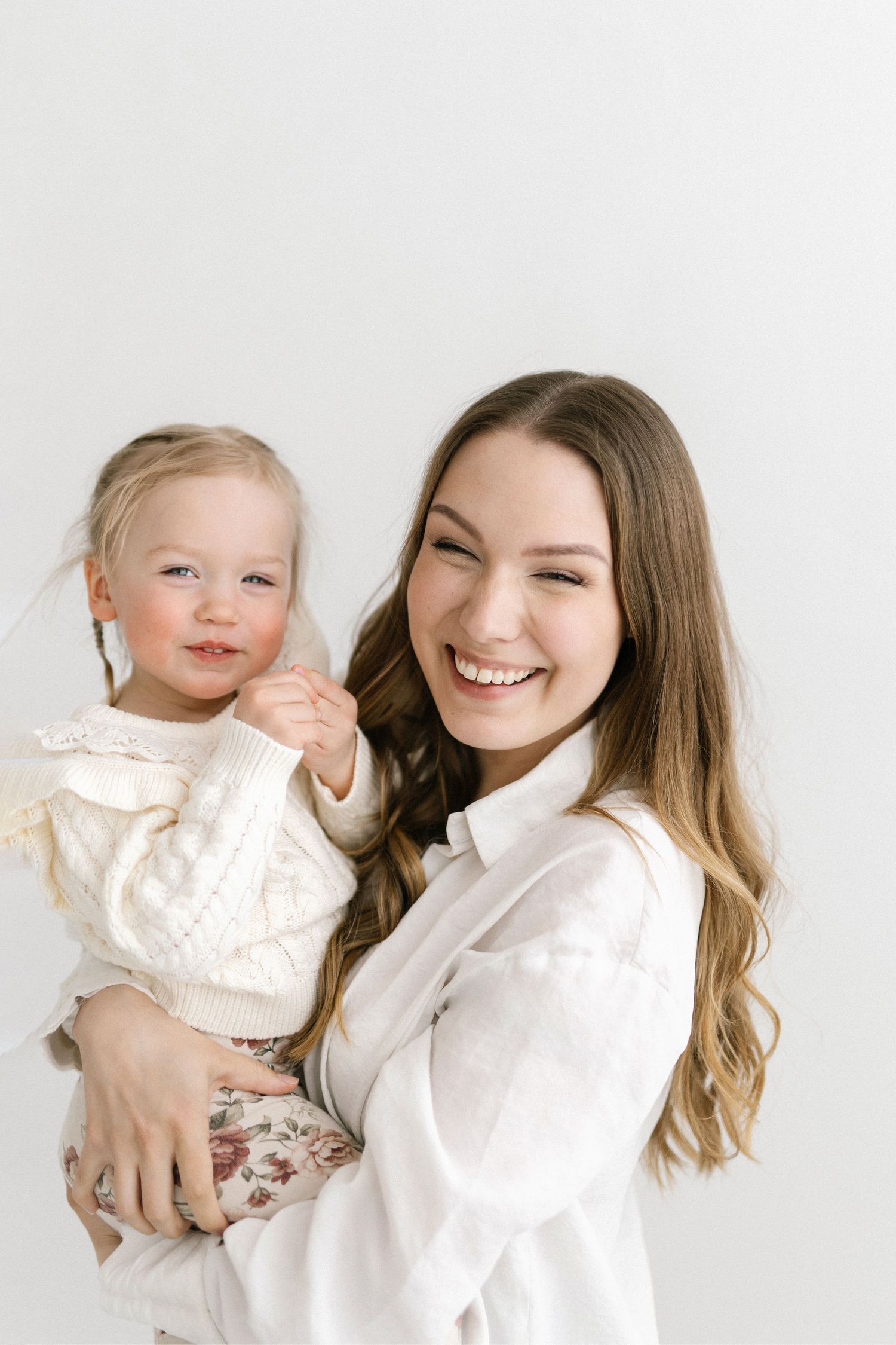 Smiling young woman in white shirt holding a happy toddler girl in a cream knit sweater against white background.
