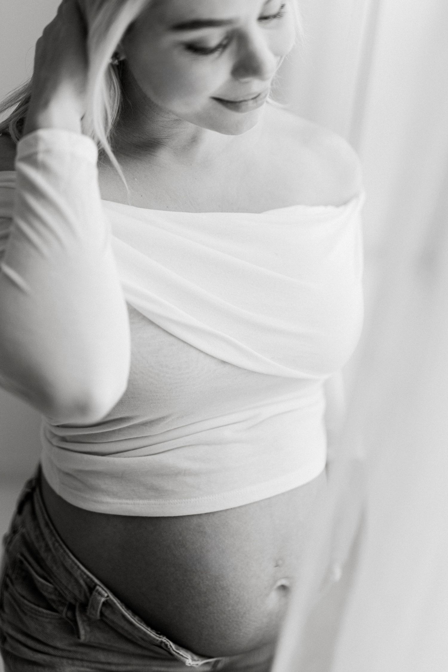 Black and white maternity photo of a pregnant woman in a white wrap top and jeans, gently cradling her baby bump.
