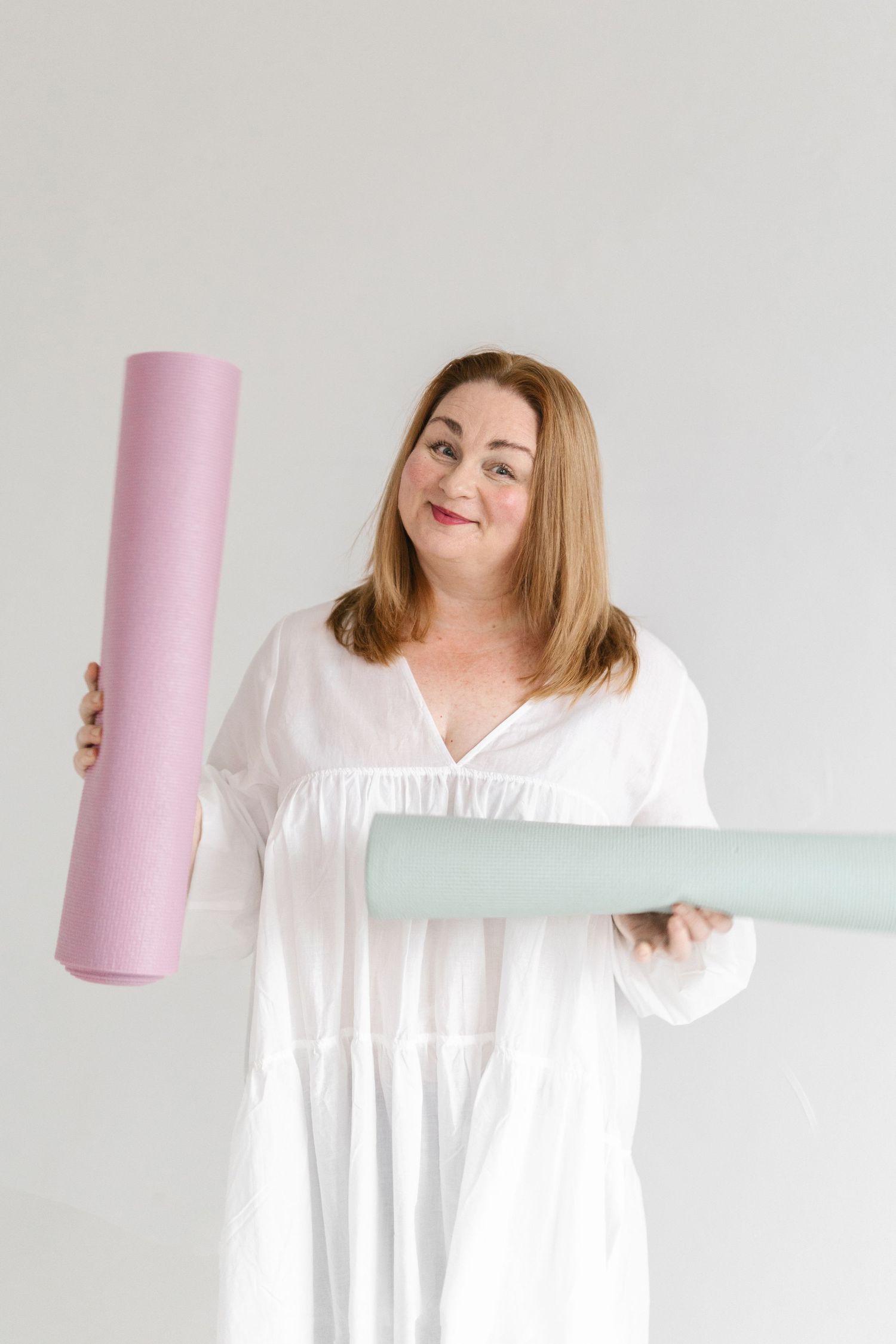 Woman in white dress holding pink and mint yoga mats against neutral background.