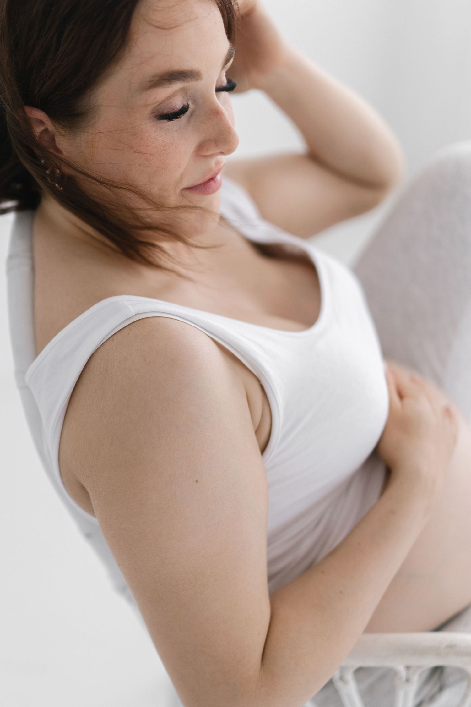 Pregnant woman in a white tank top and light leggings sits gently holding her belly with one hand behind her head in soft natural light.