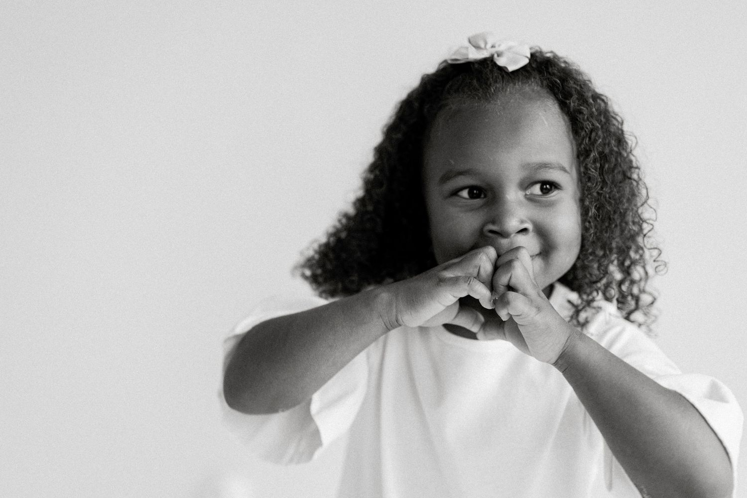 Black and white portrait of a smiling young girl with curly hair and a bow, hands clasped near her chin as she looks to the side.