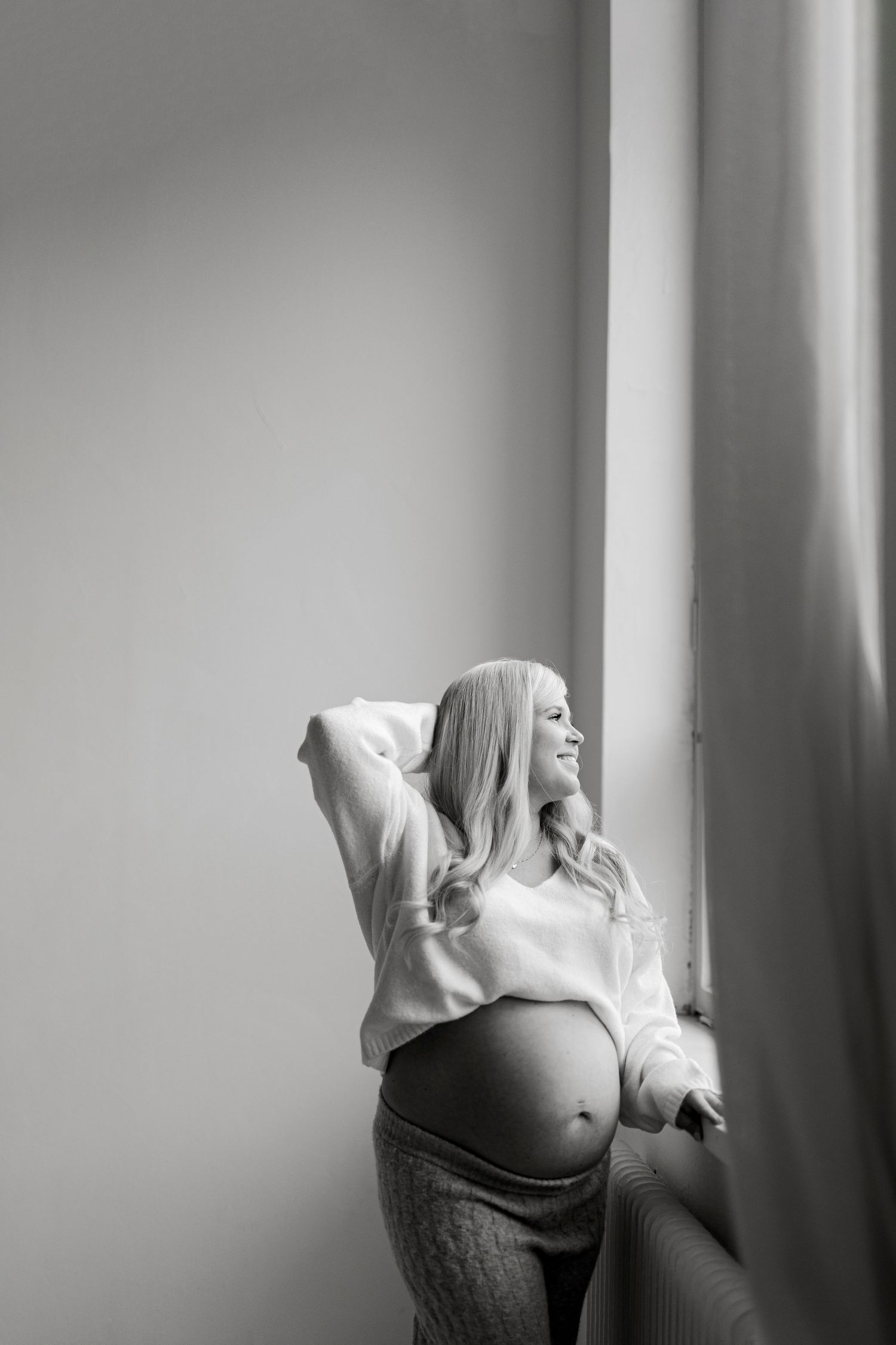Black and white maternity portrait of a smiling pregnant woman in a cropped sweater standing by a bright window with her arm raised.