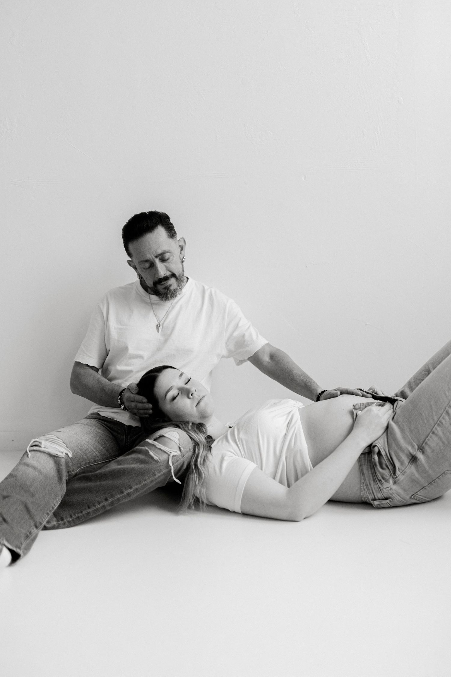 Black and white maternity photo of a couple sitting on the floor, woman resting her head on mans lap.