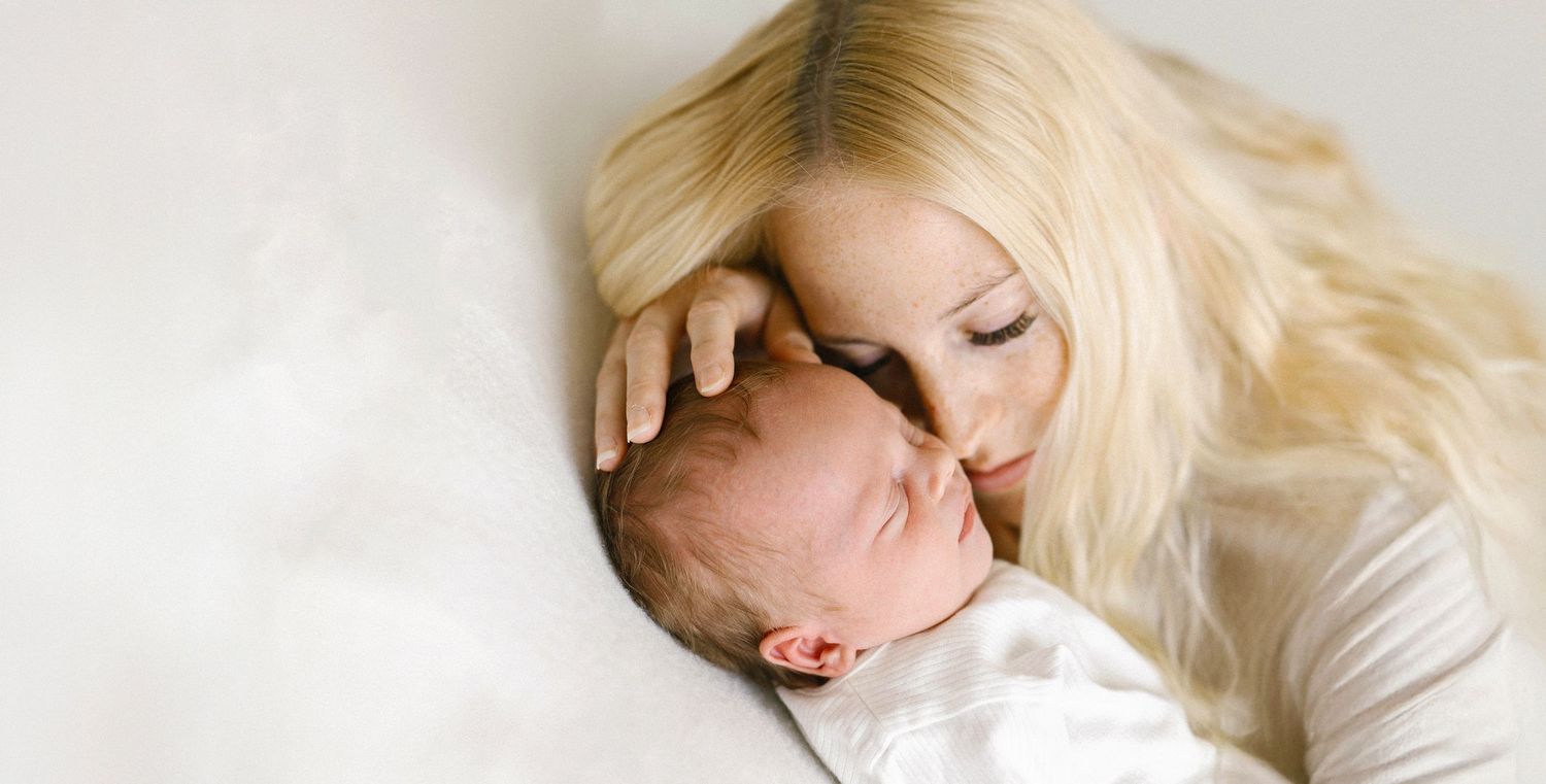 Blonde mother tenderly kissing her newborn baby wrapped in white swaddle while lying together on soft white bedding.