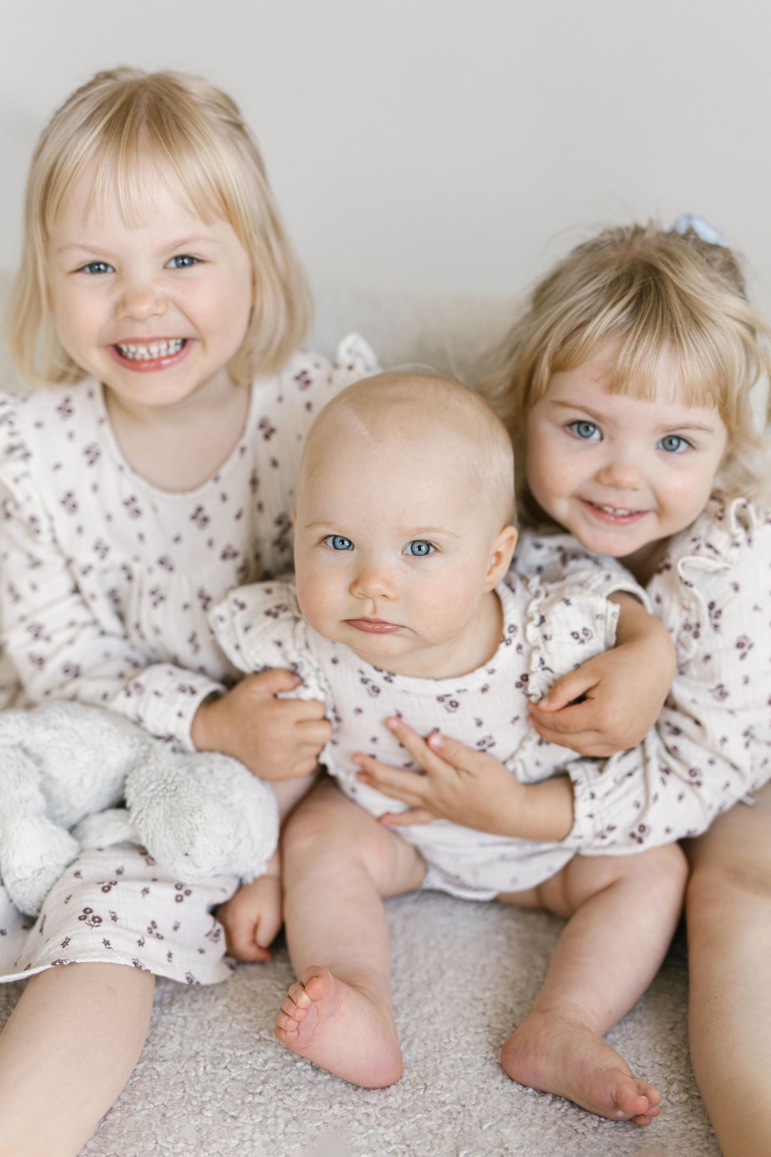Three blonde blue-eyed siblings in matching floral outfits pose together, two toddler girls holding a smiling baby.