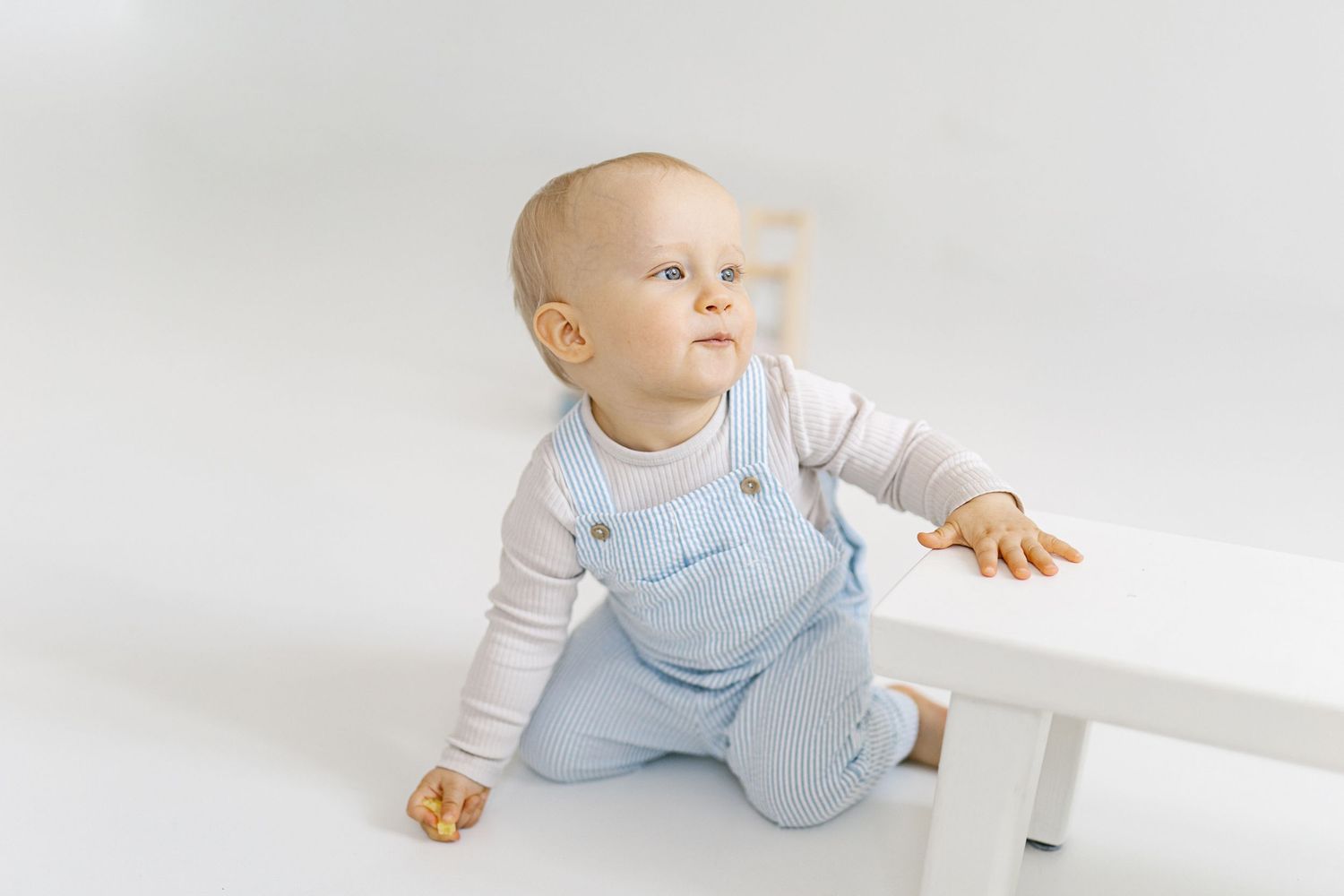 A cute baby in light blue overalls sits on a white floor, leaning on a small white table, looking up curiously.