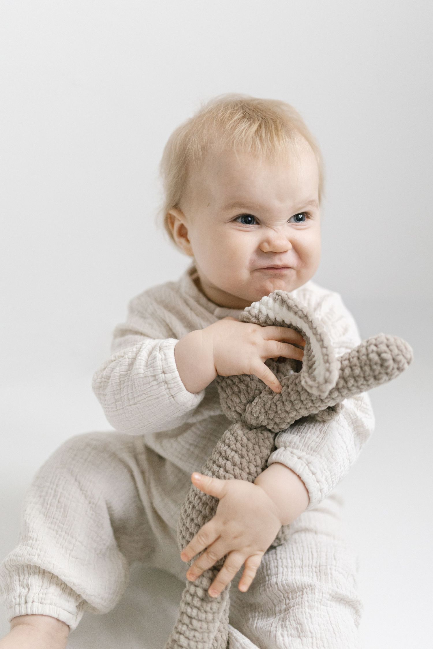 Baby in white sweater holding and playing with gray knit stuffed toy.