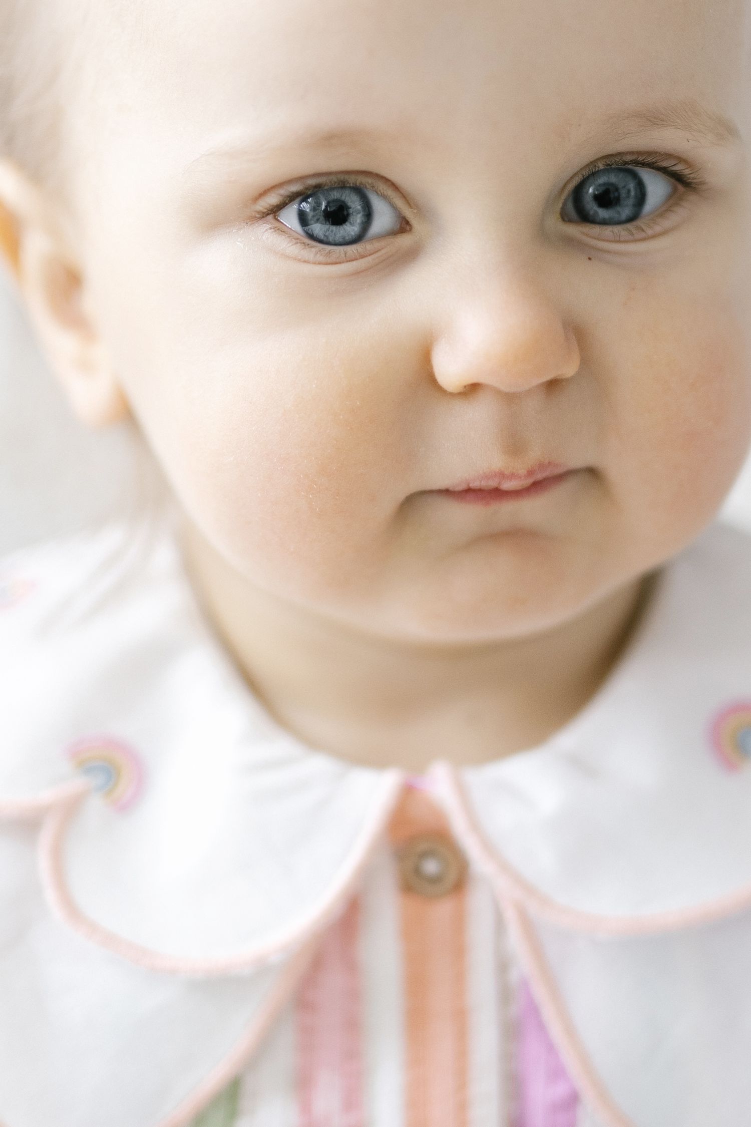 Close-up portrait of an infant with bright blue eyes wearing a pink and white outfit against a light background.