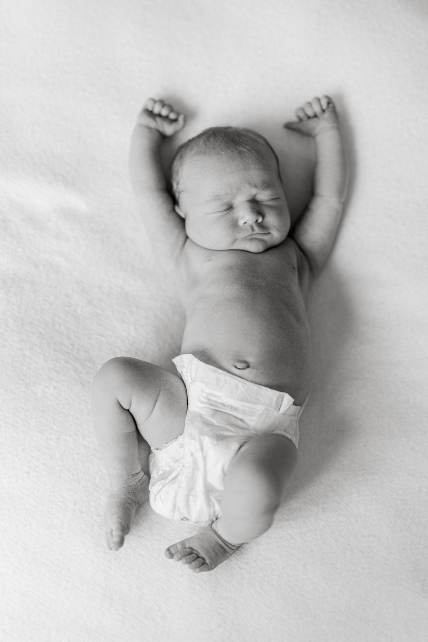 Black and white photo of a sleeping newborn baby in a diaper lying on a white blanket with arms raised.