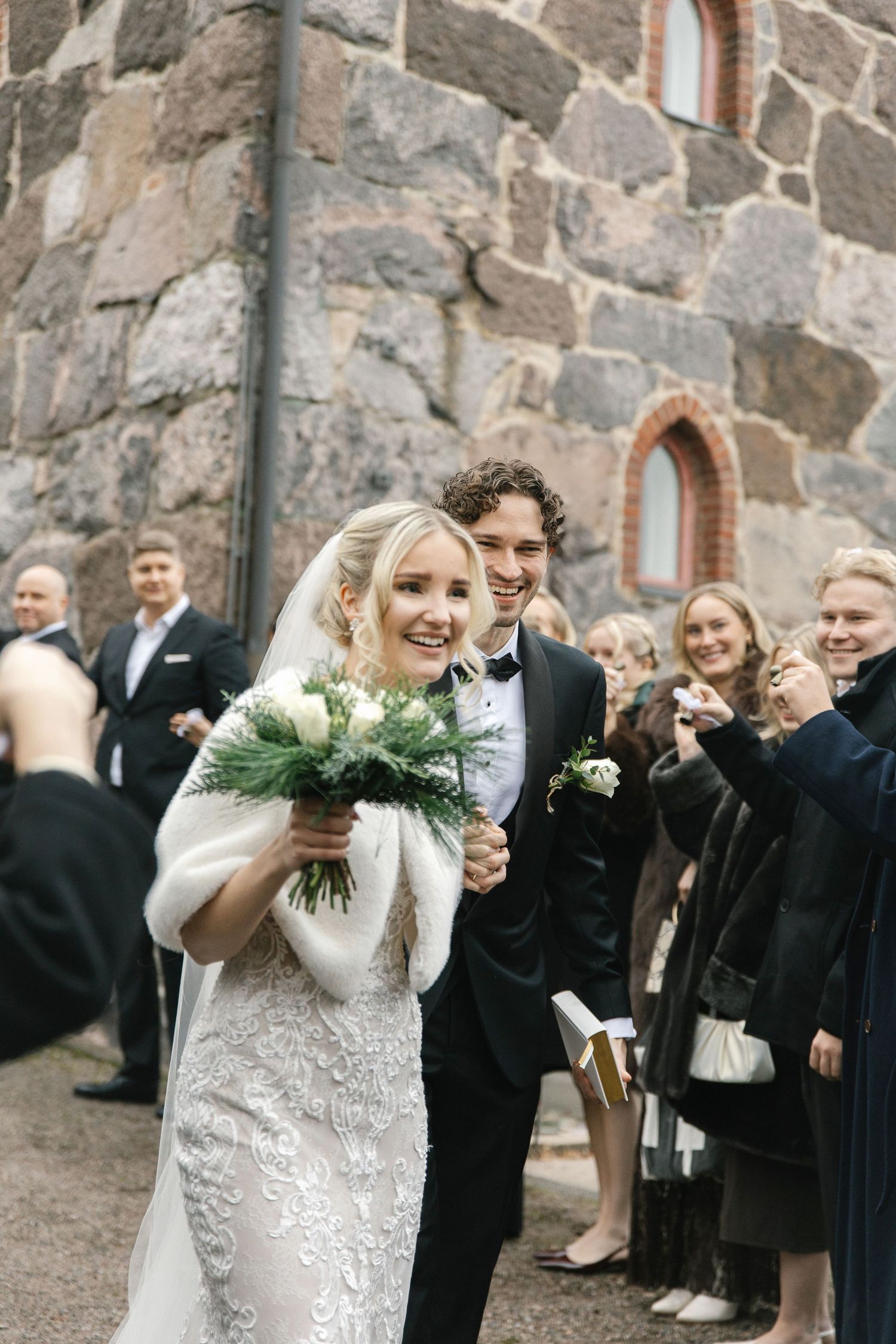 Wedding party celebrating outdoors near stone building with evergreen bouquet and formal attire.