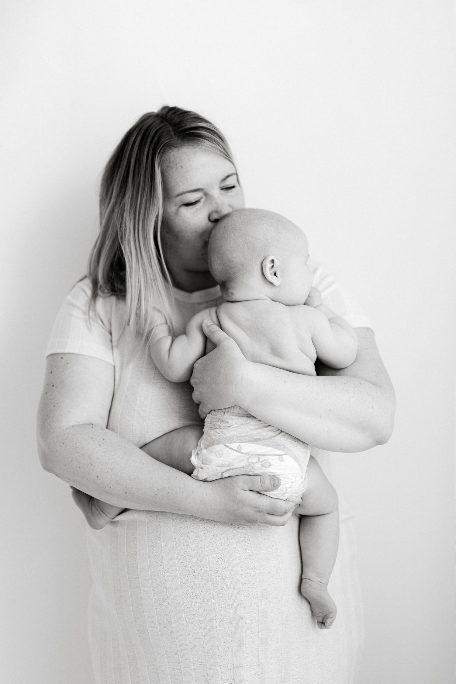 Black and white photo of a mother tenderly kissing her newborn baby while cradling them in her arms.