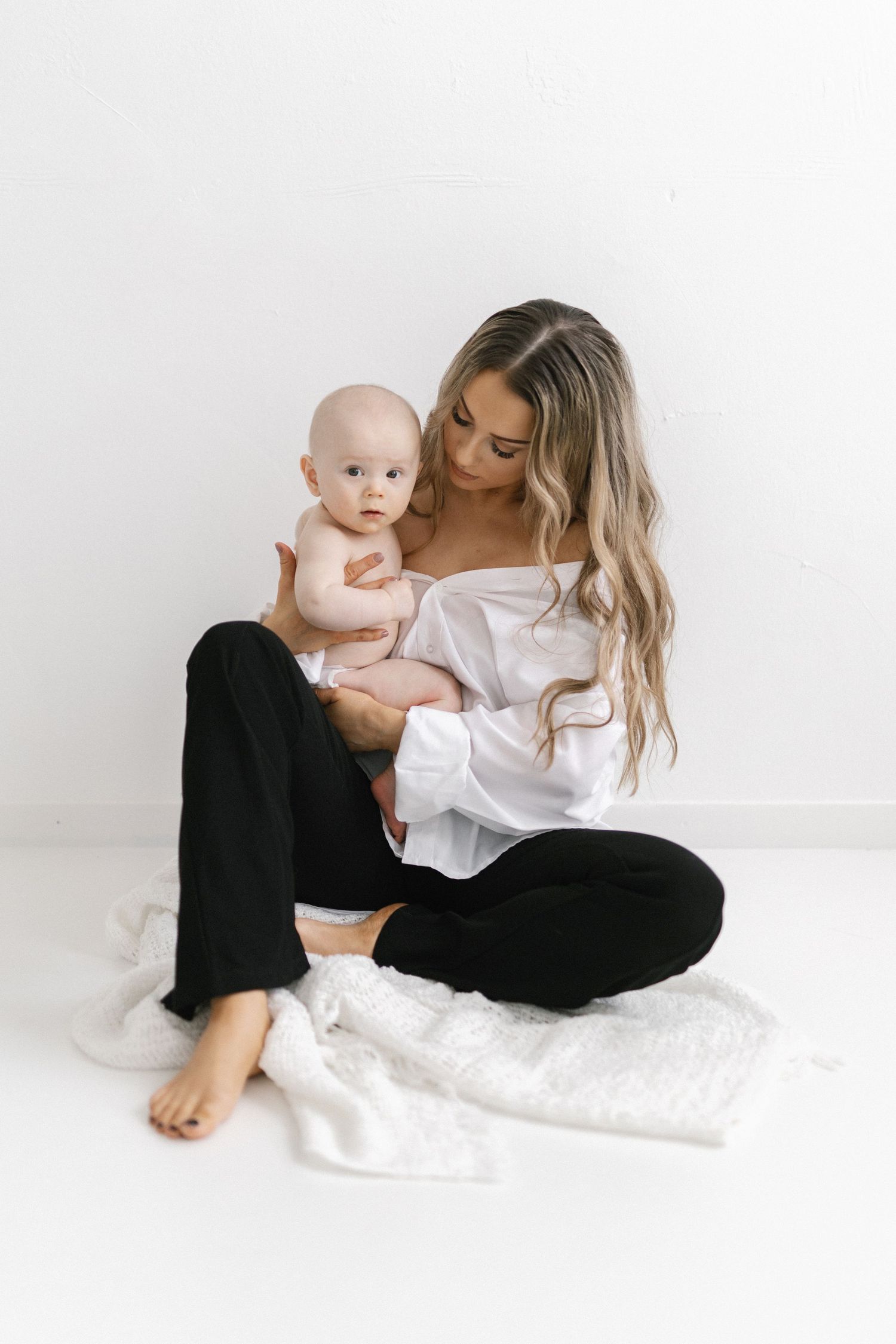 A young mother with long blonde hair sits cross-legged on the floor holding her baby, wearing a white top and black pants.