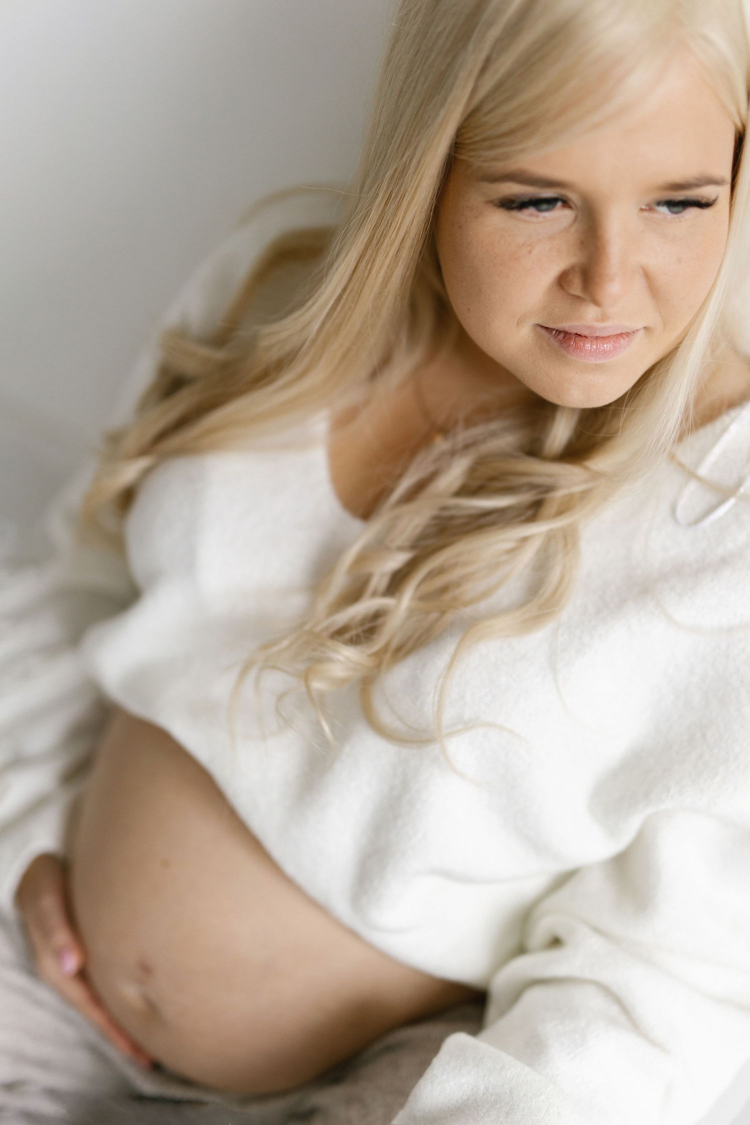 Pregnant woman with long wavy blonde hair in a cozy white knit sweater gently resting her hand on her bare baby bump.