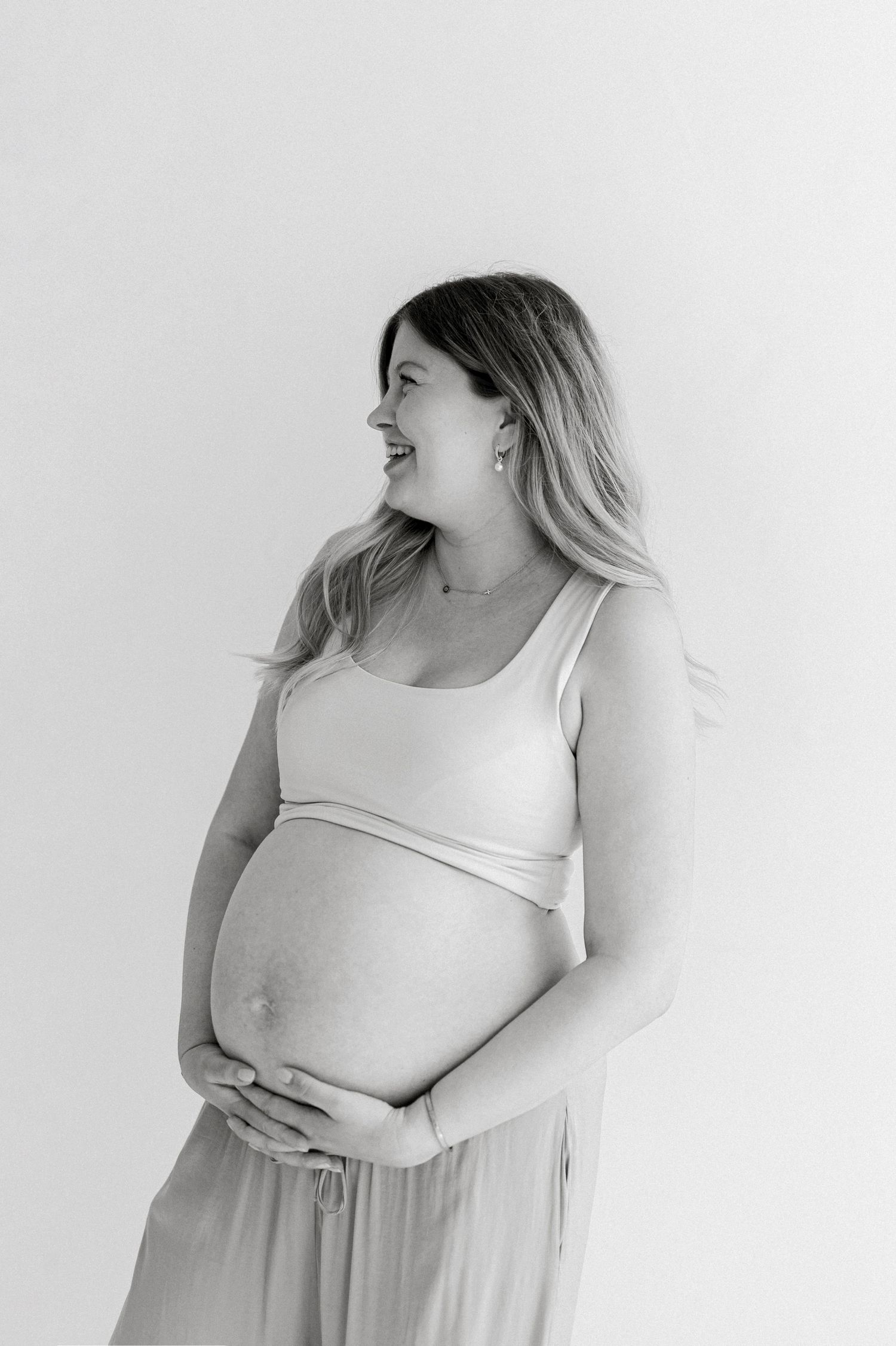 Black and white maternity portrait of a smiling pregnant woman with long hair cradling her bare belly against a plain studio backdrop.