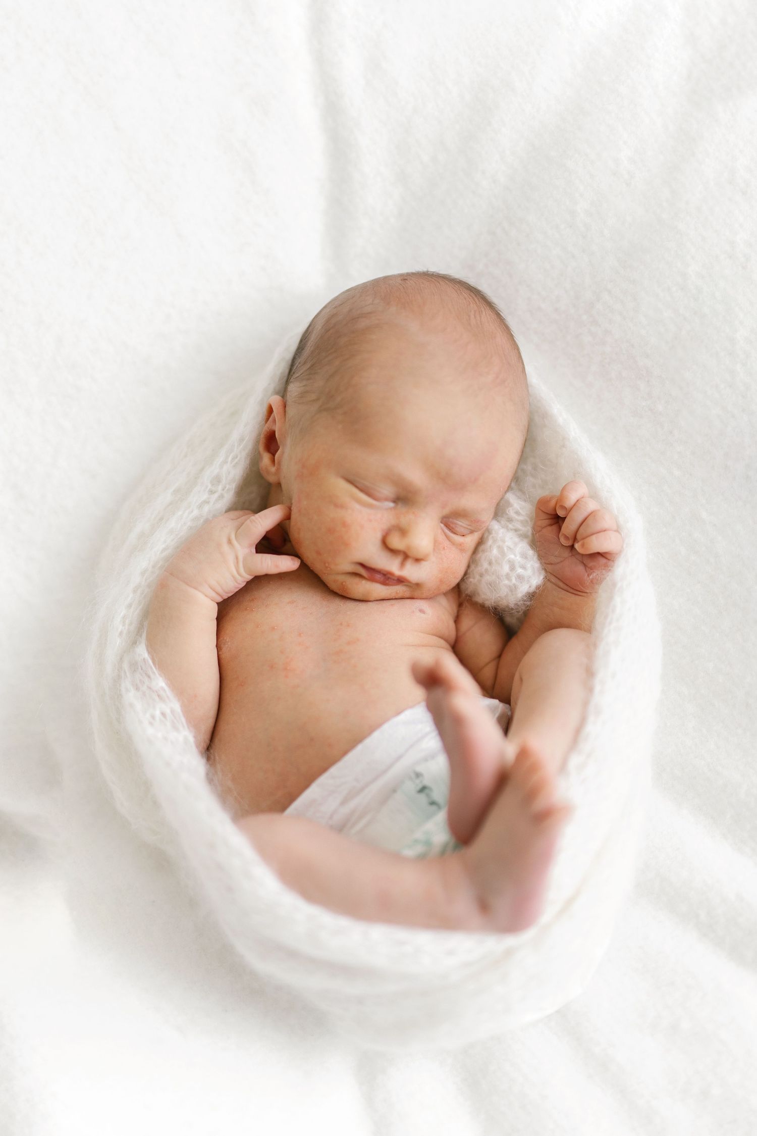 Sleeping newborn baby wrapped in a soft white knit blanket on a white background, with tiny hands and feet in a peaceful newborn photo.