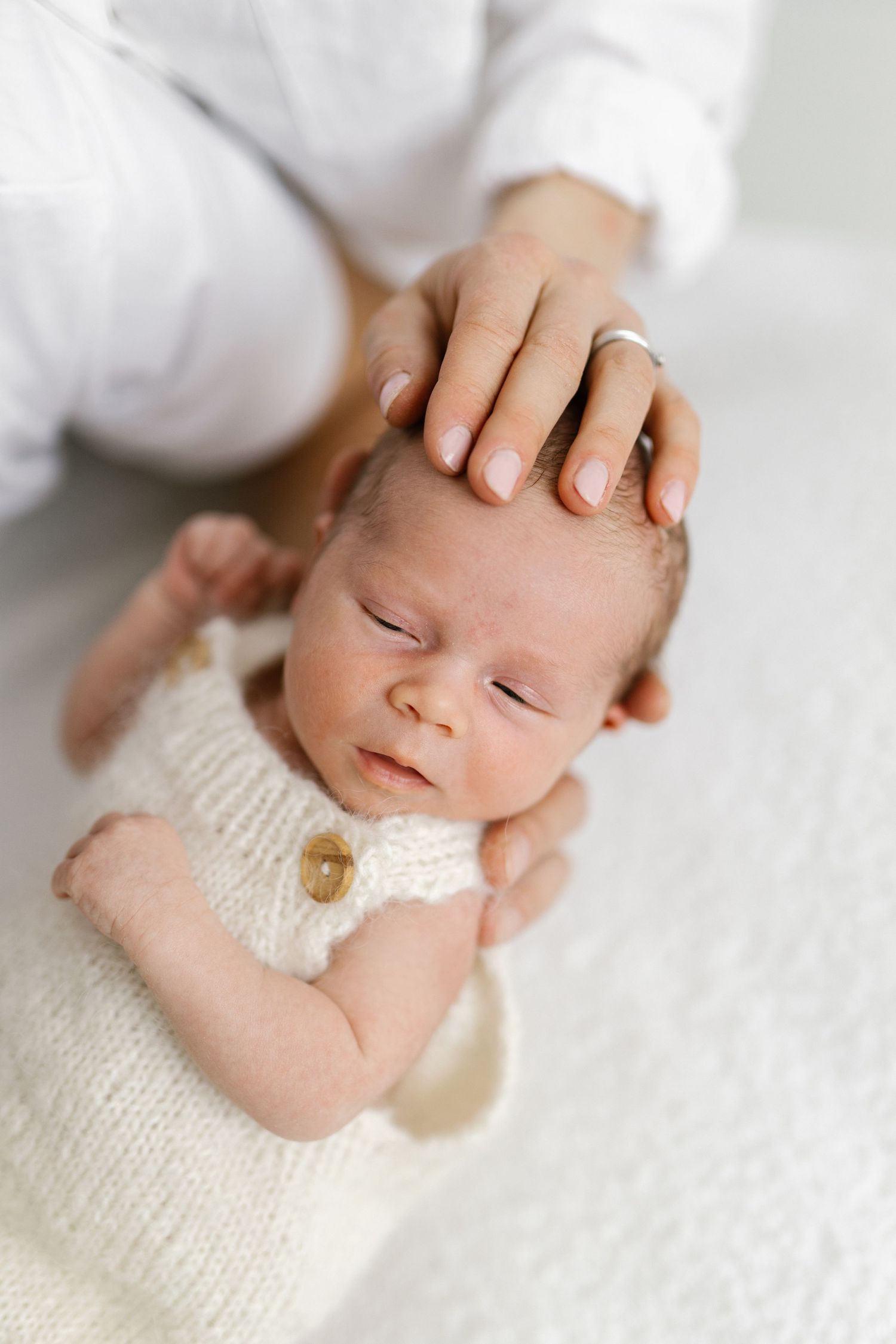 Newborn baby sleeping peacefully in cream knit romper while parent gently cradles head with ringed hand.