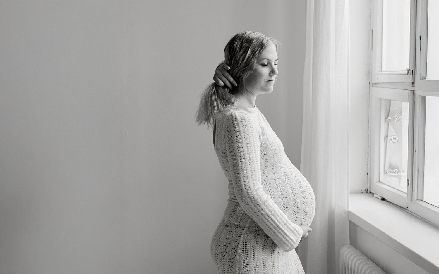 Black and white photo of a pregnant woman in a white dress gazing out a window in a serene, elegant maternity portrait.