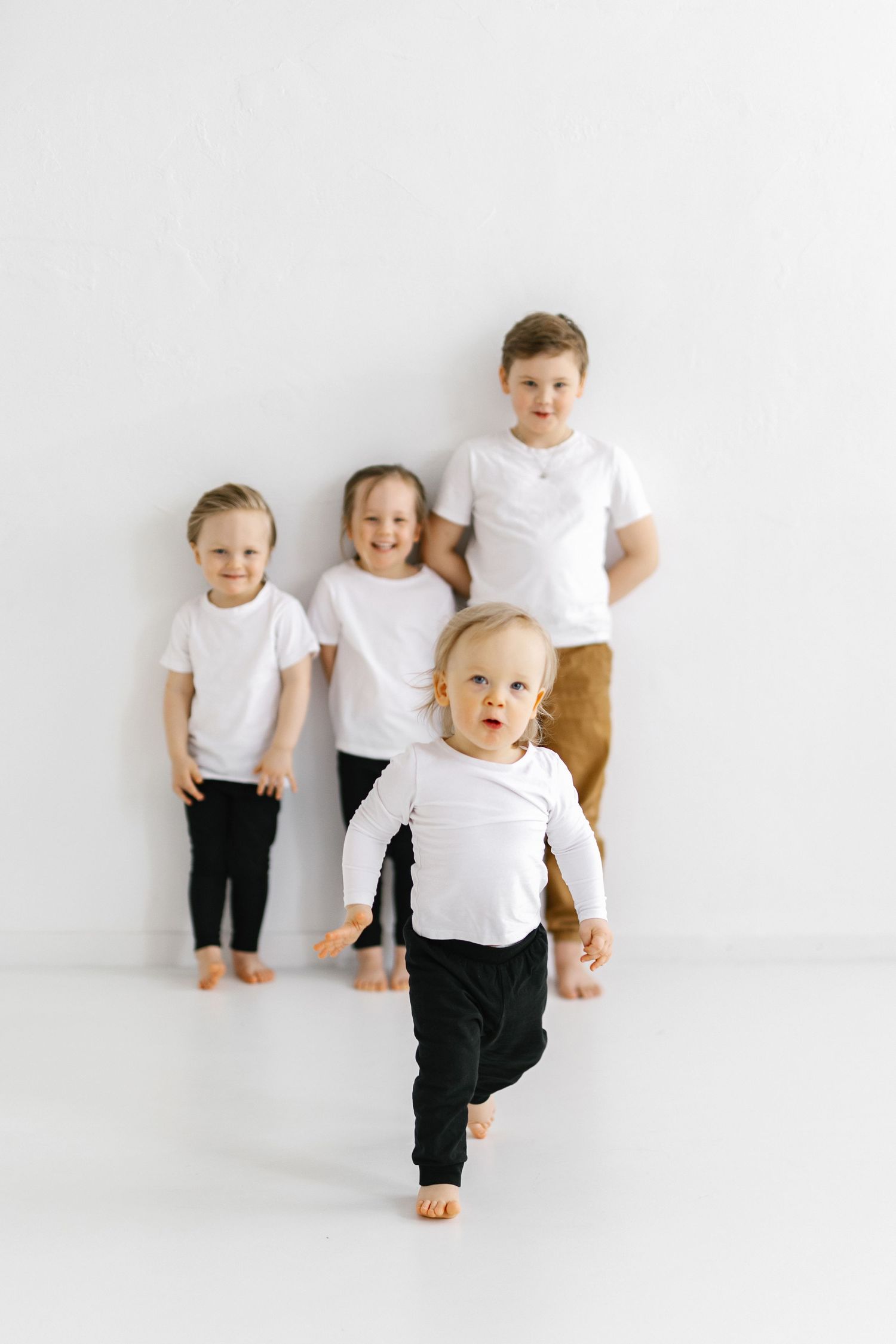 Children in matching white t-shirts and black pants play and pose together against a white backdrop.