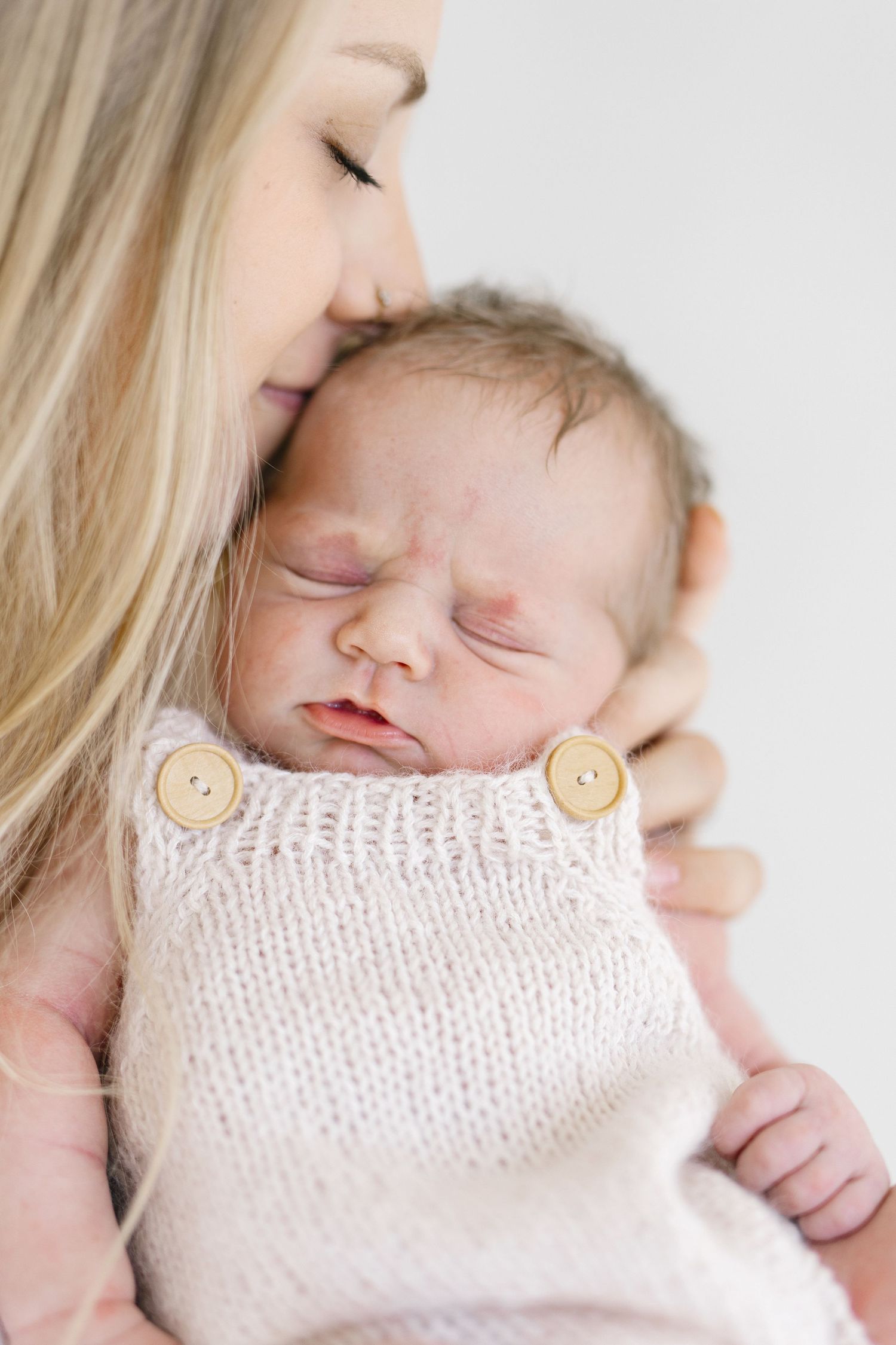 A mother with blonde hair gently kisses her sleeping newborn baby dressed in a white knit romper with wooden buttons.
