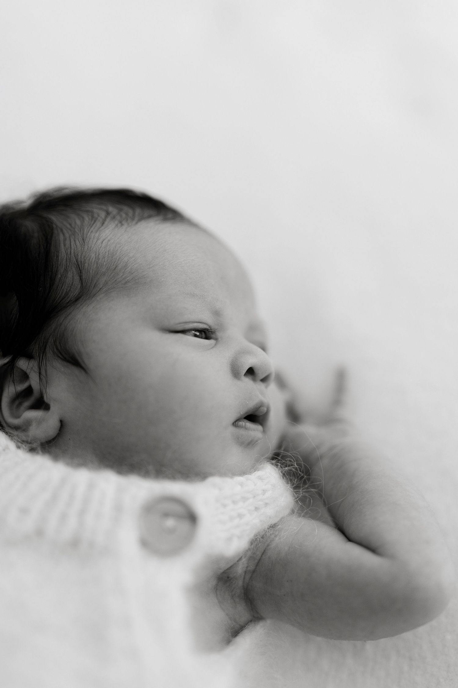 Black and white portrait of a sleeping newborn baby wearing a knit romper with buttons, lying on a soft white blanket.