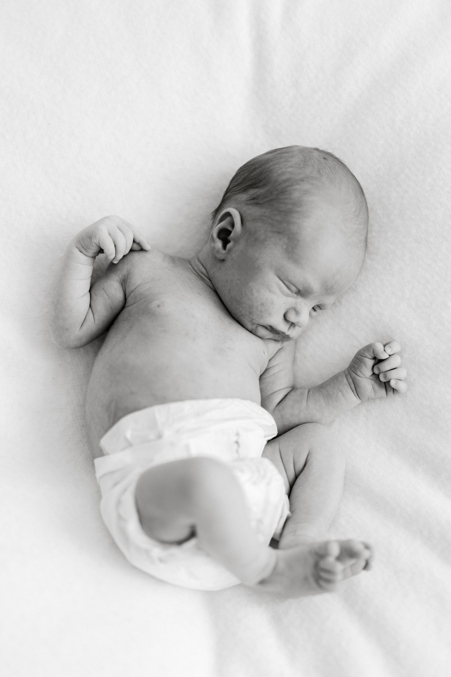 Black and white photo of a sleeping newborn baby in a diaper lying on a soft white blanket.