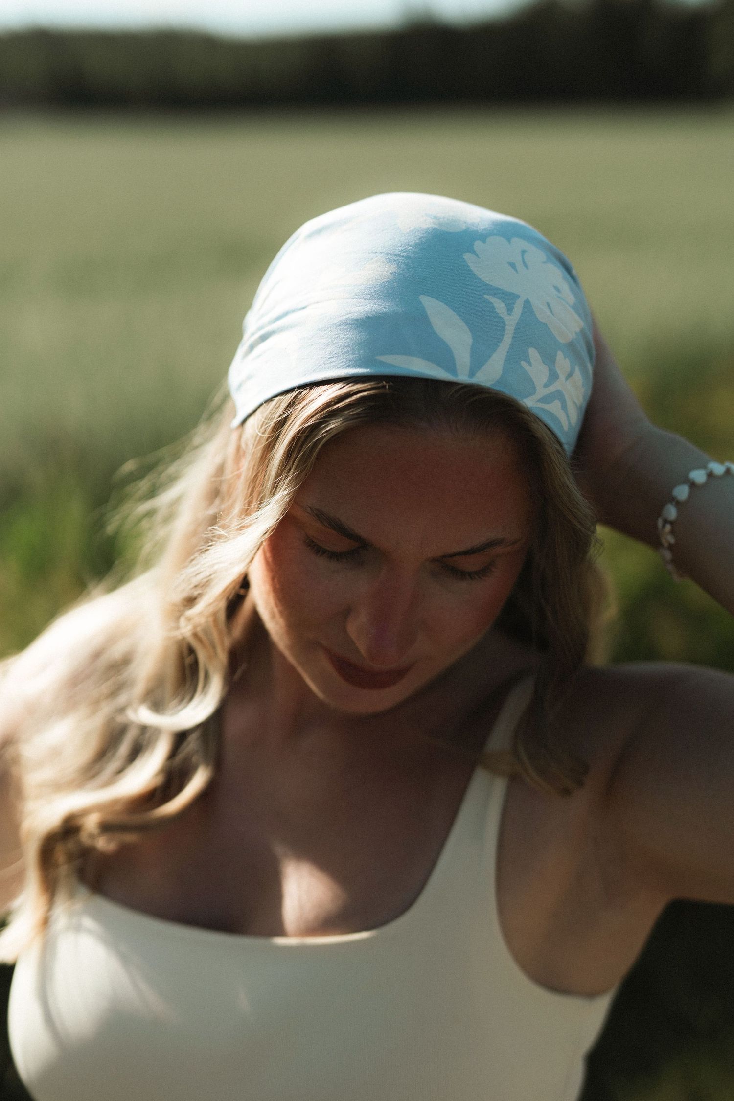 Woman wearing a light blue floral headband outdoors in a sunny green field, with blonde wavy hair and a white top.