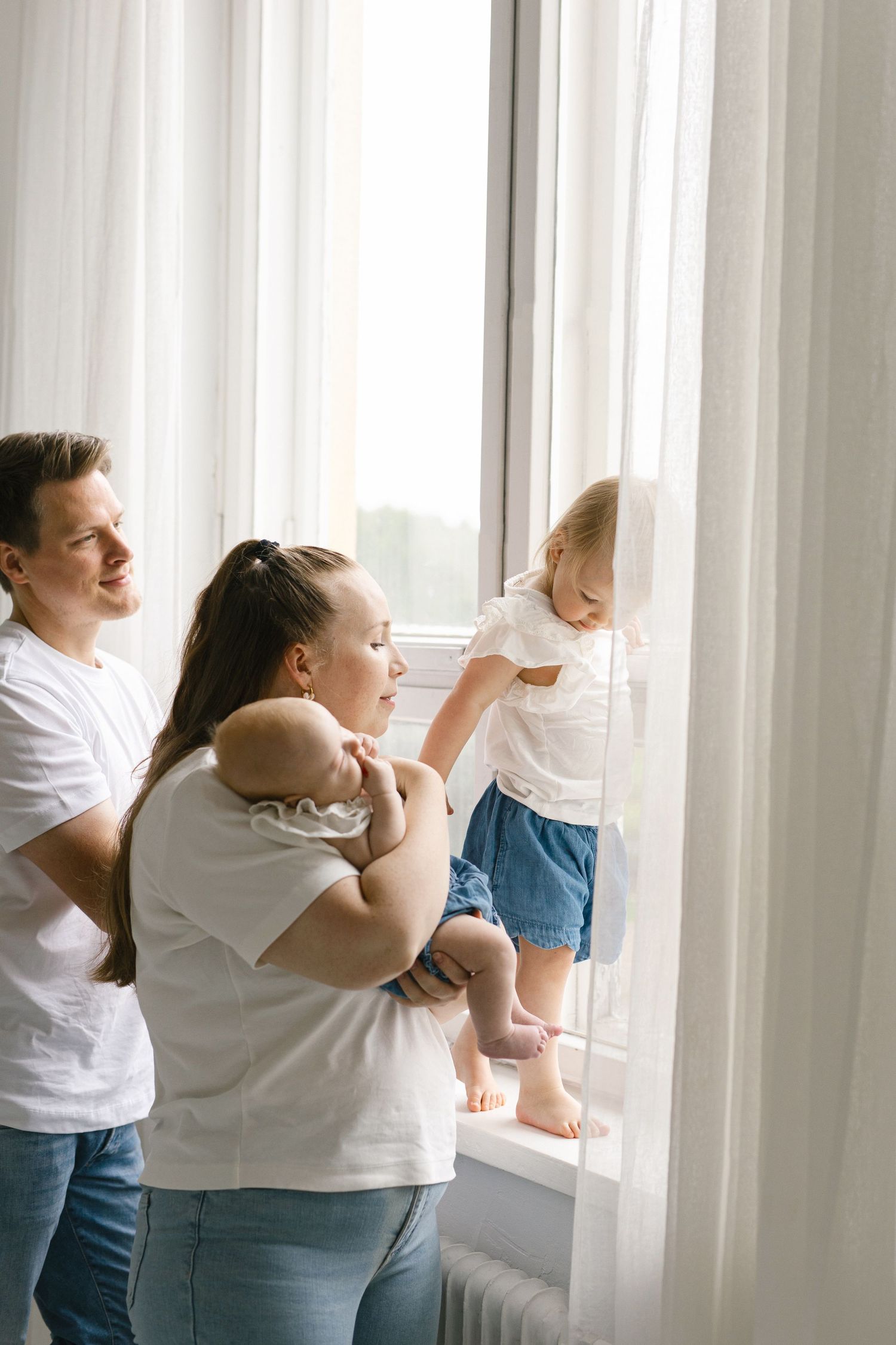 A young family with a baby and toddler standing by a bright window, dressed in white shirts and jeans.