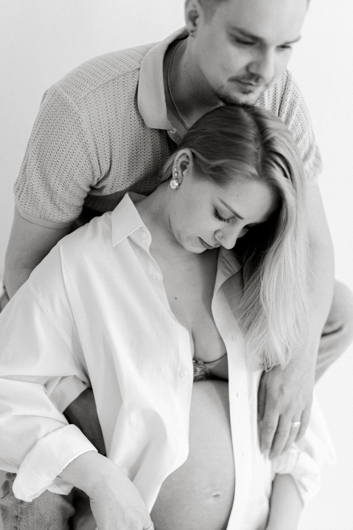 Black and white photo of pregnant woman in open white shirt embraced by her partner from behind.
