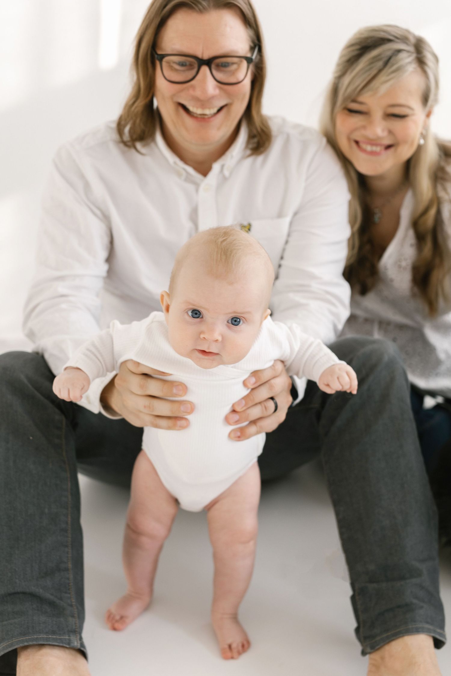 Happy parents in white shirts holding their blue-eyed baby in a white onesie in a bright studio setting.