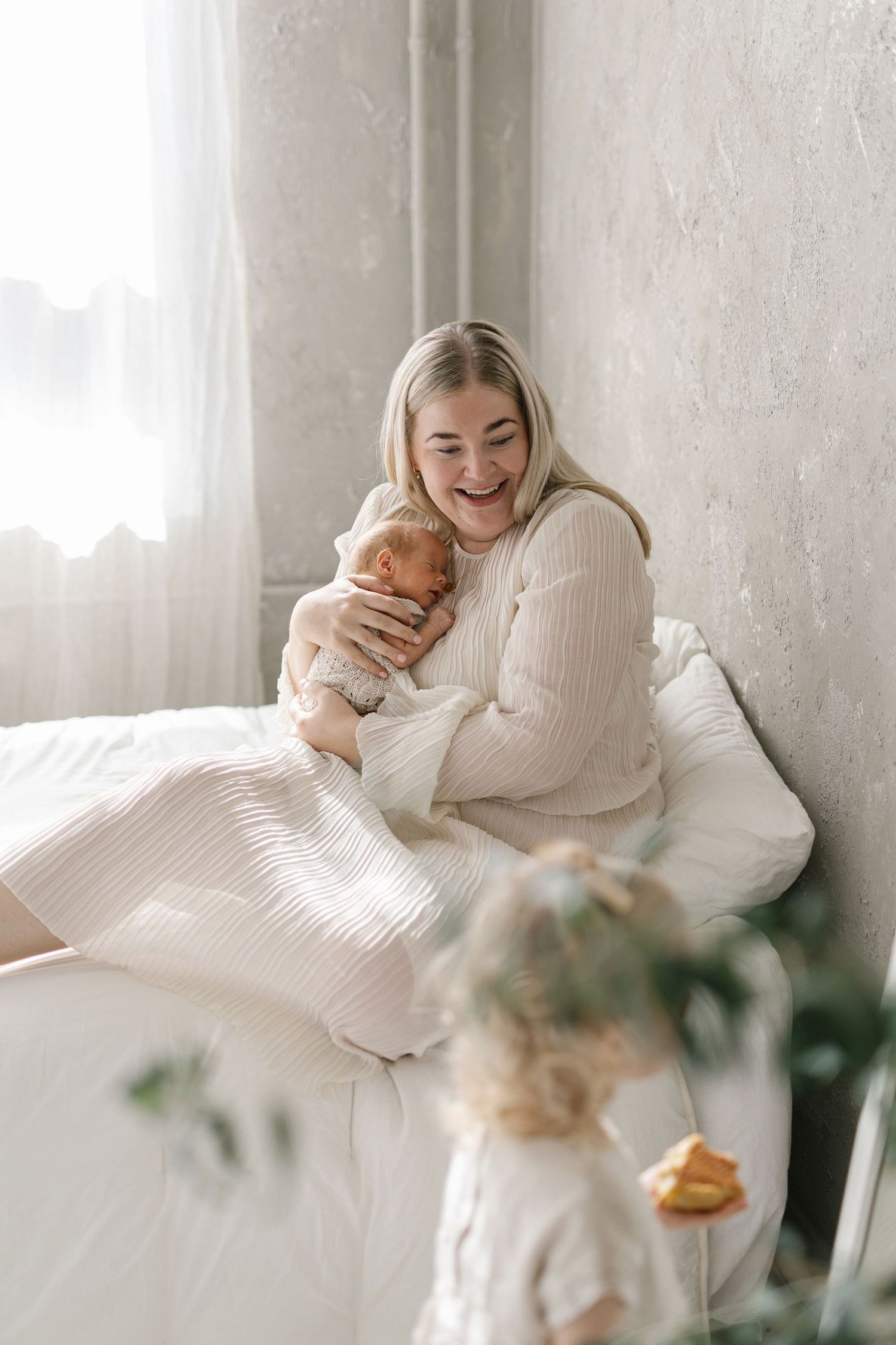 A joyful mother holds her newborn baby while sitting on a bed, with a toddler standing nearby in a bright room.