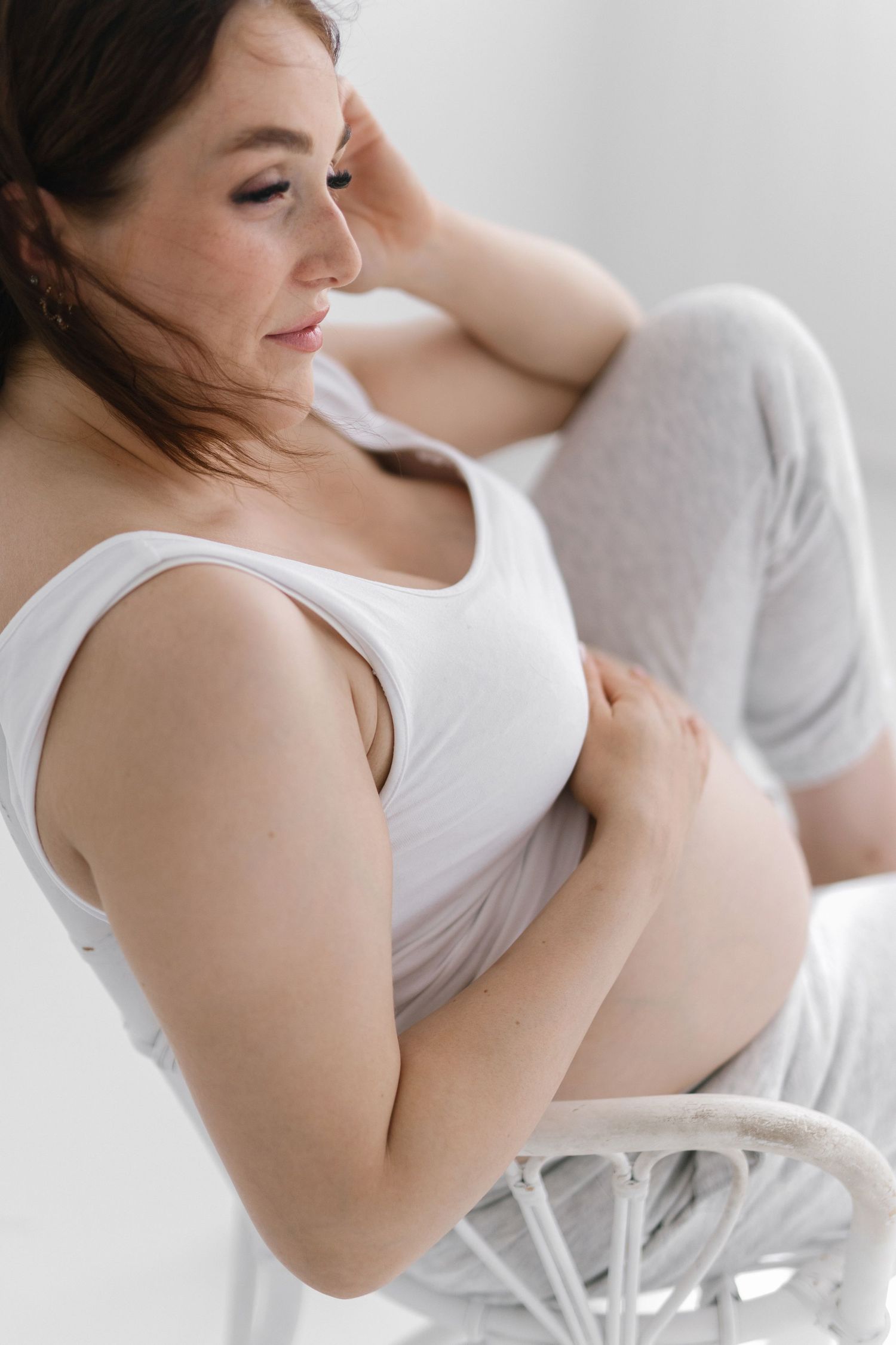 Pregnant woman in white tank top sitting in wicker chair, cradling her baby bump with a serene expression.