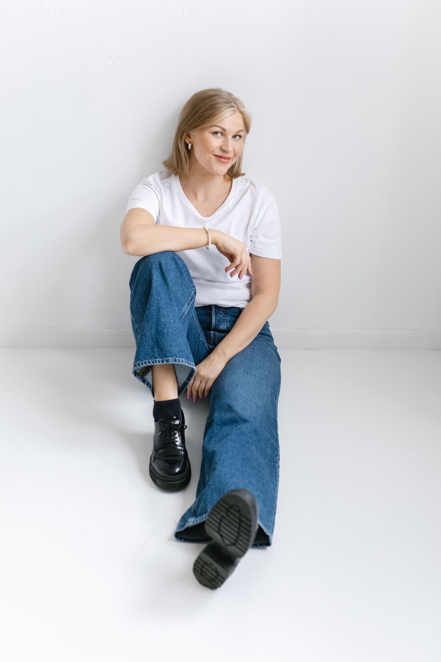 Person in casual white t-shirt and wide-leg jeans sits against white wall wearing black boots.