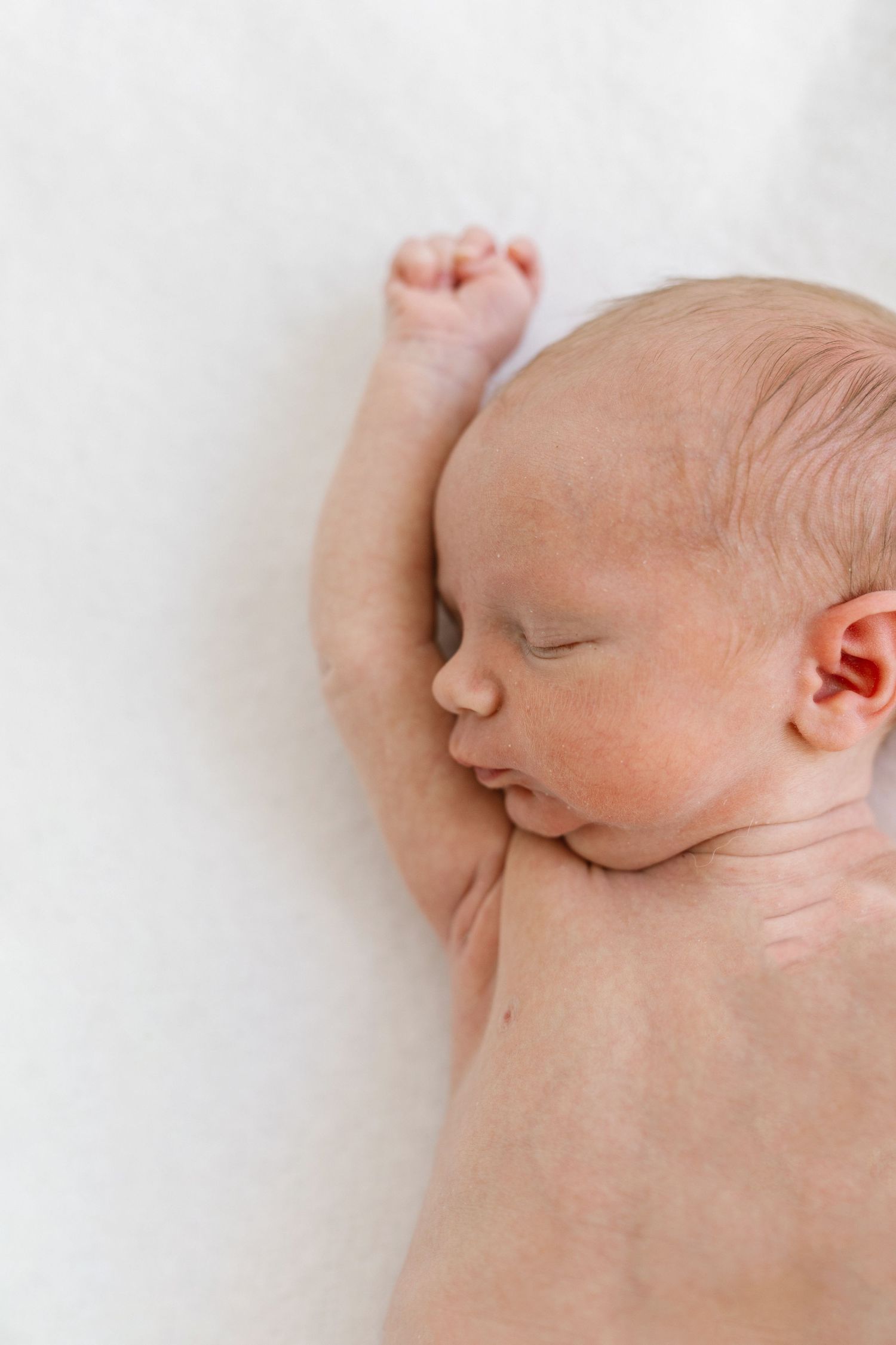Peaceful newborn baby sleeping on a white blanket with arm raised, captured in a soft natural light portrait.
