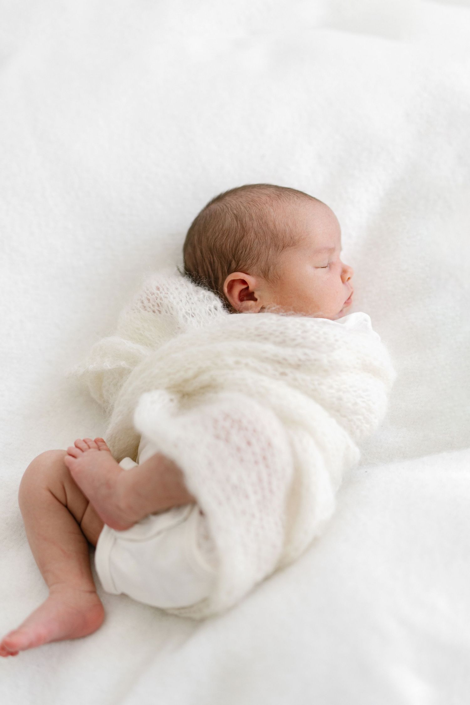 Peaceful newborn baby sleeping wrapped in soft white knit blanket on white bedding.