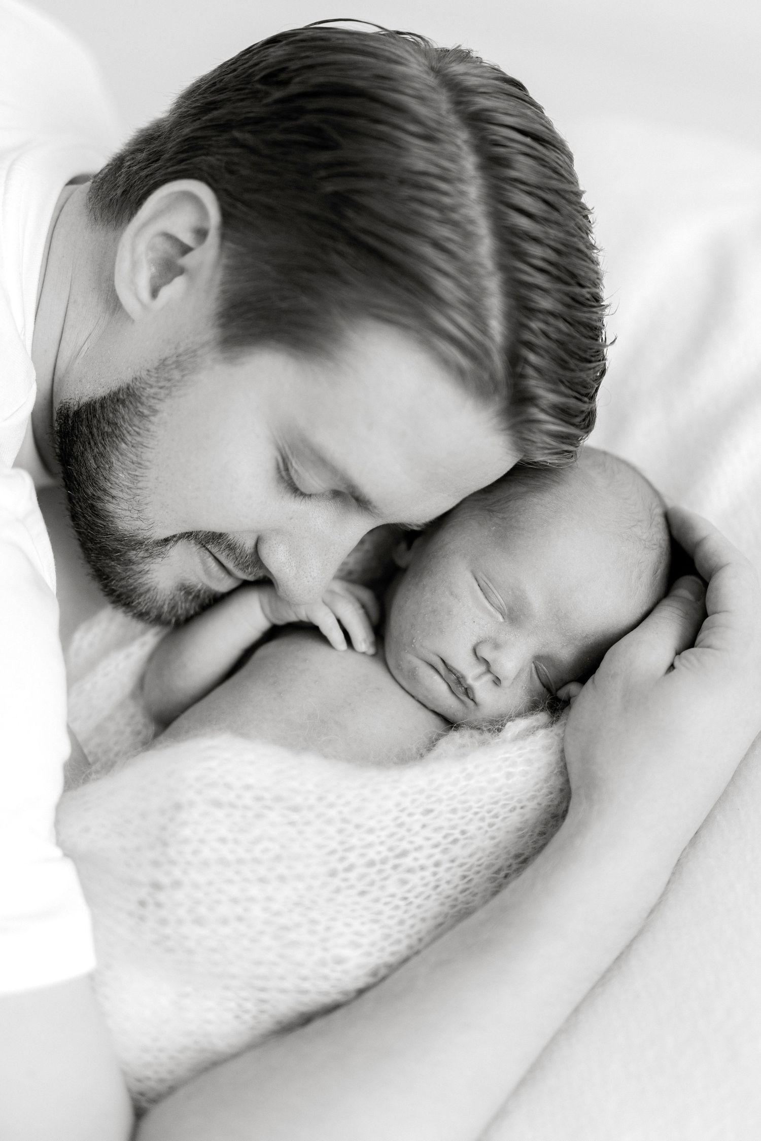 Black and white photo of a father tenderly kissing his sleeping newborn baby wrapped in a soft blanket.