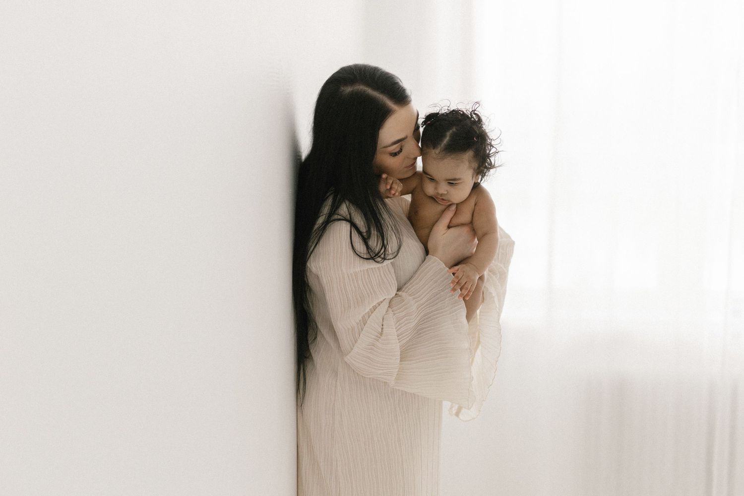A mother tenderly holds her baby close, both dressed in white, sharing a gentle moment in a bright, minimalist room.