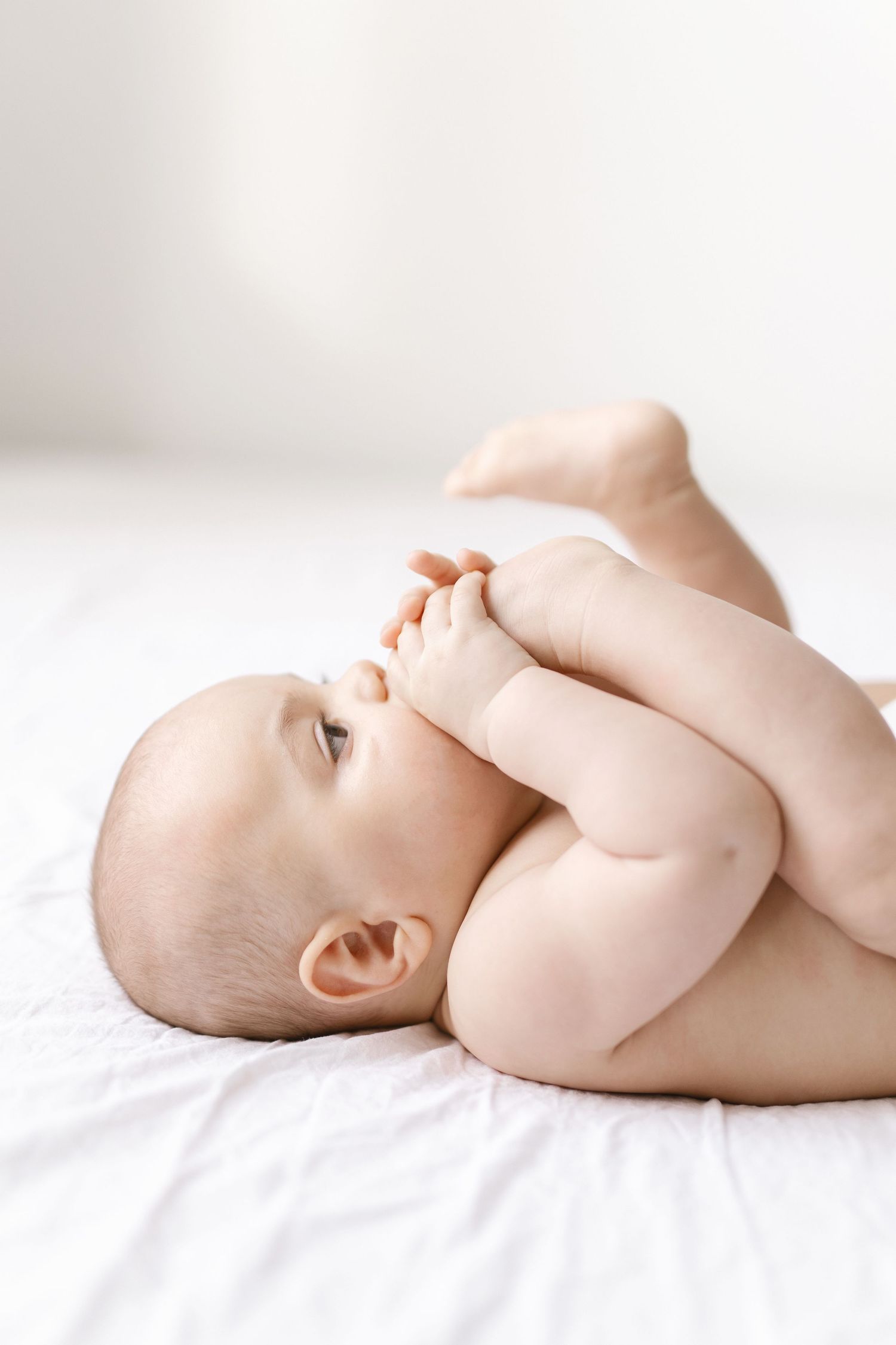 Naked baby lying on white bed, playfully holding feet up while looking to the side in a bright, airy room.