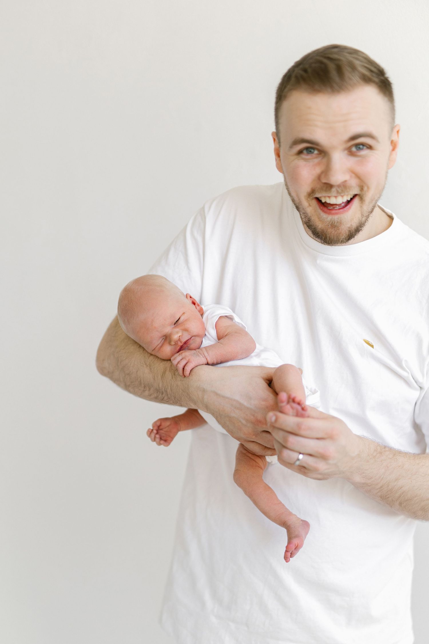 Excited young father in white t-shirt proudly holds sleeping newborn baby against white background.