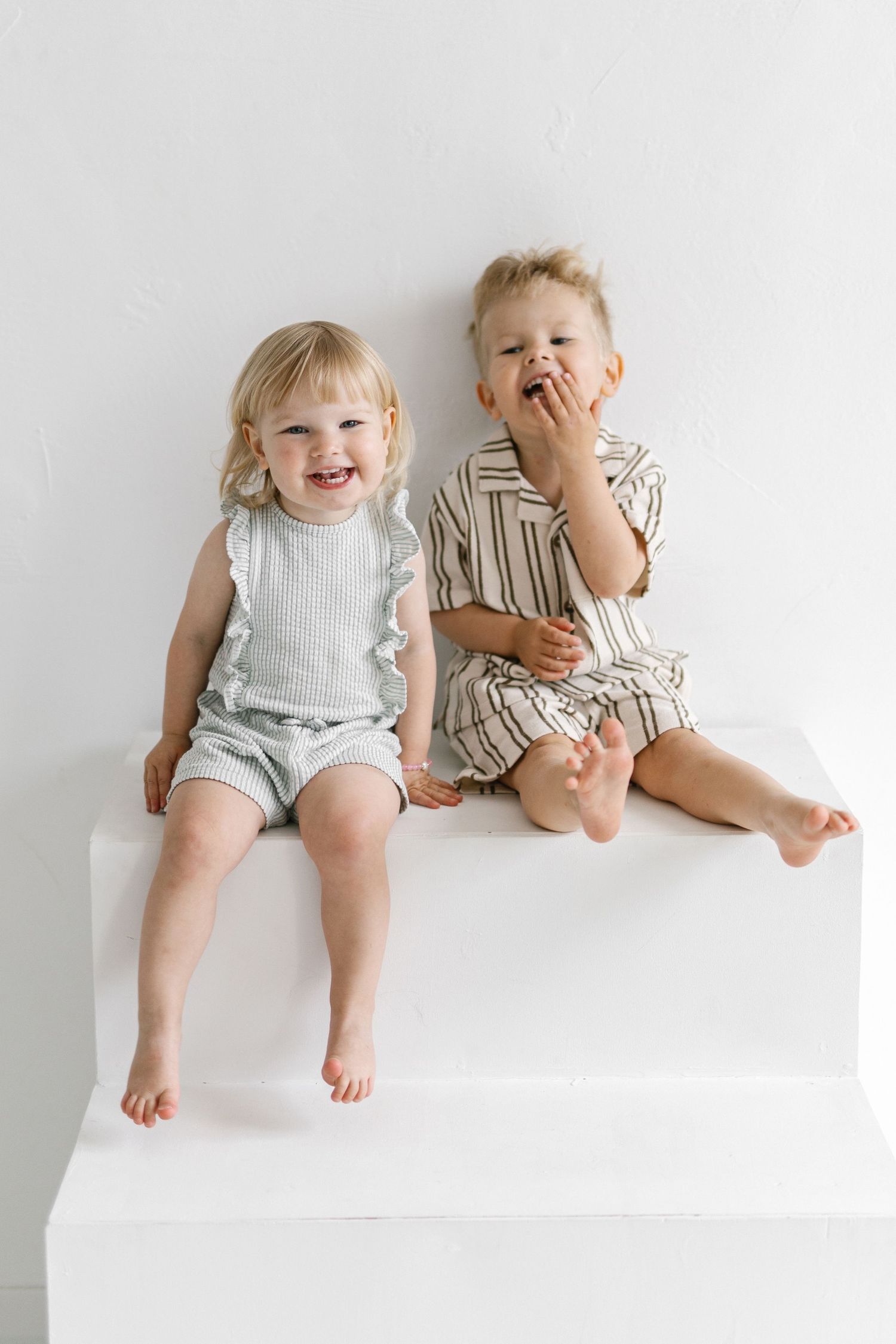Two happy toddlers sitting barefoot on a white step, wearing light summer outfits, smiling and laughing together.