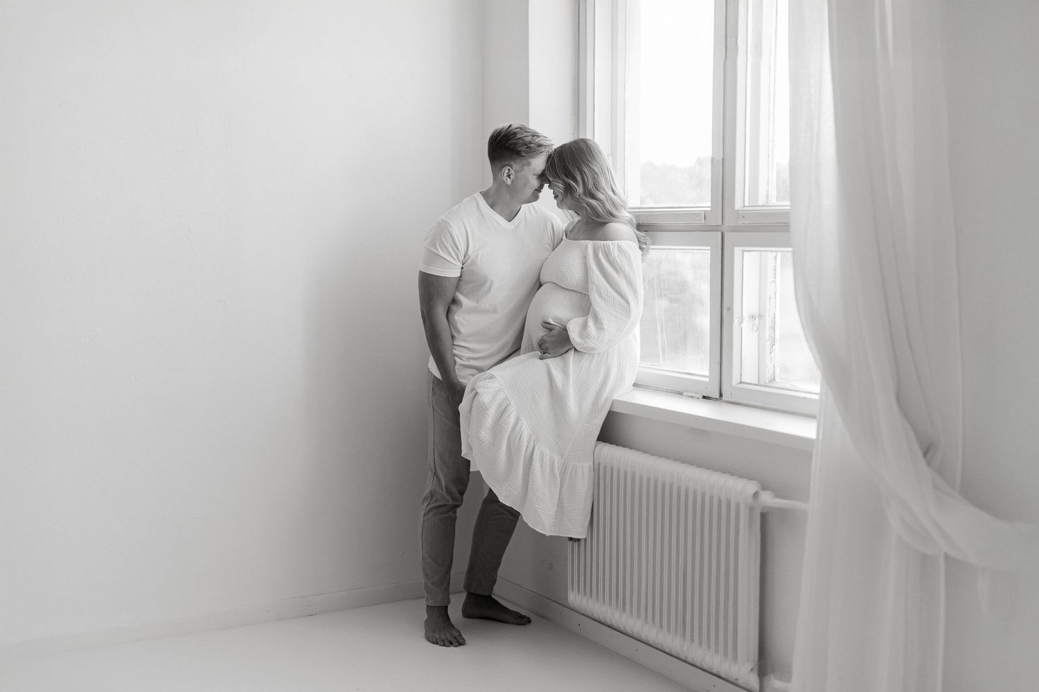 Black and white photo of a romantic couple embracing near a bright window in a minimalist white room.