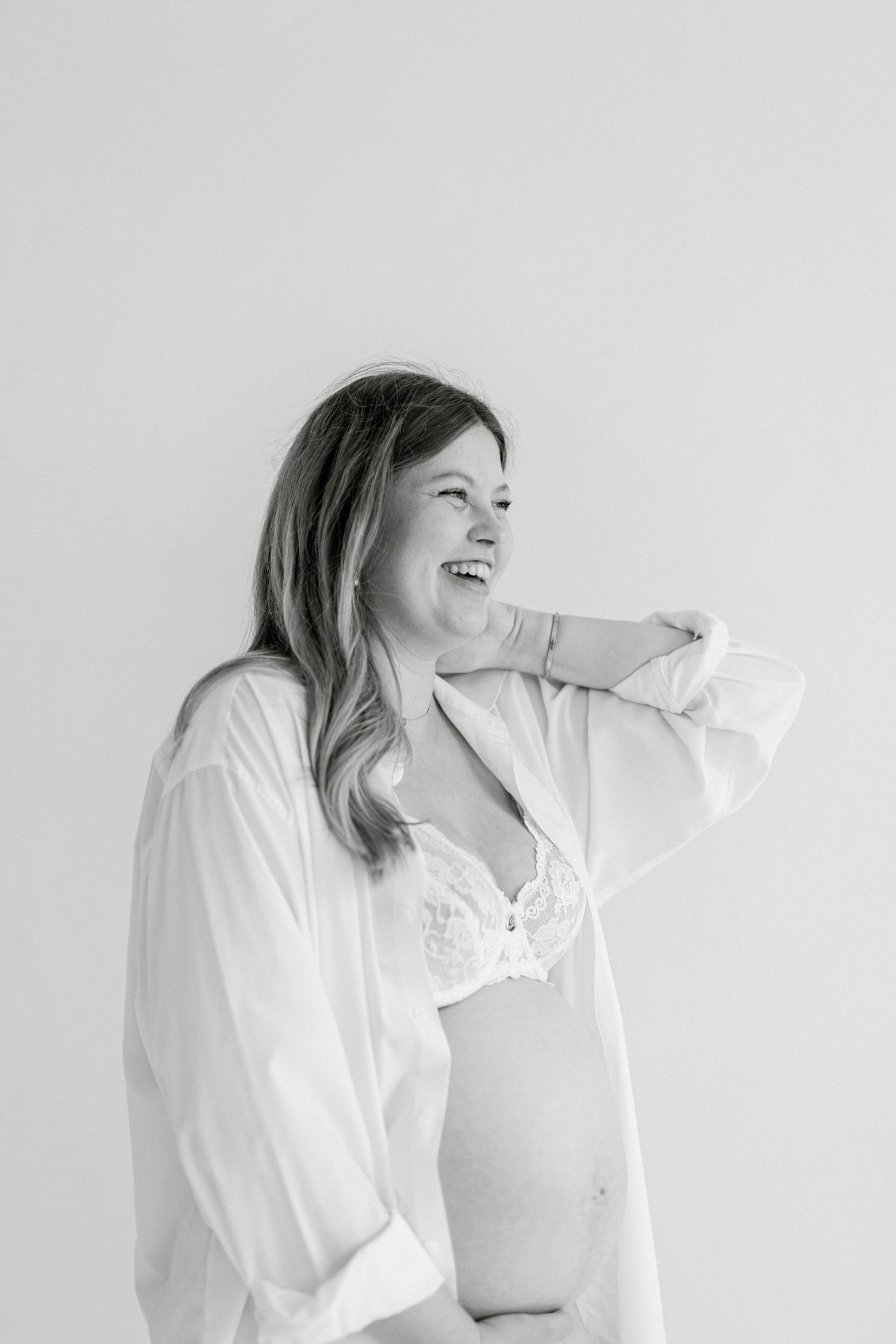 Black and white photo of a laughing pregnant woman in an open white shirt and lace bra, holding her hair back.