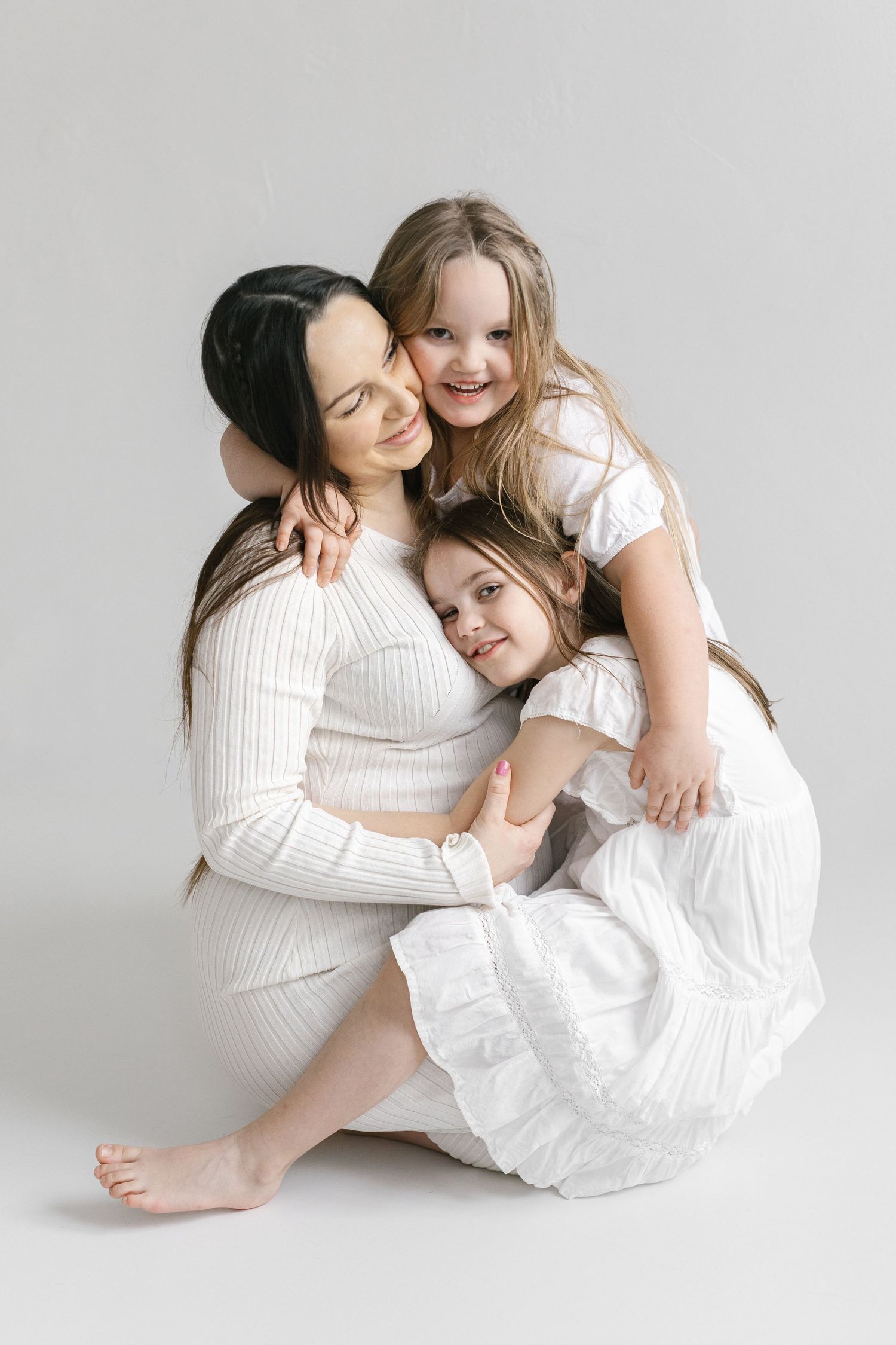 Mother and children in white clothing sharing intimate moments on light background.