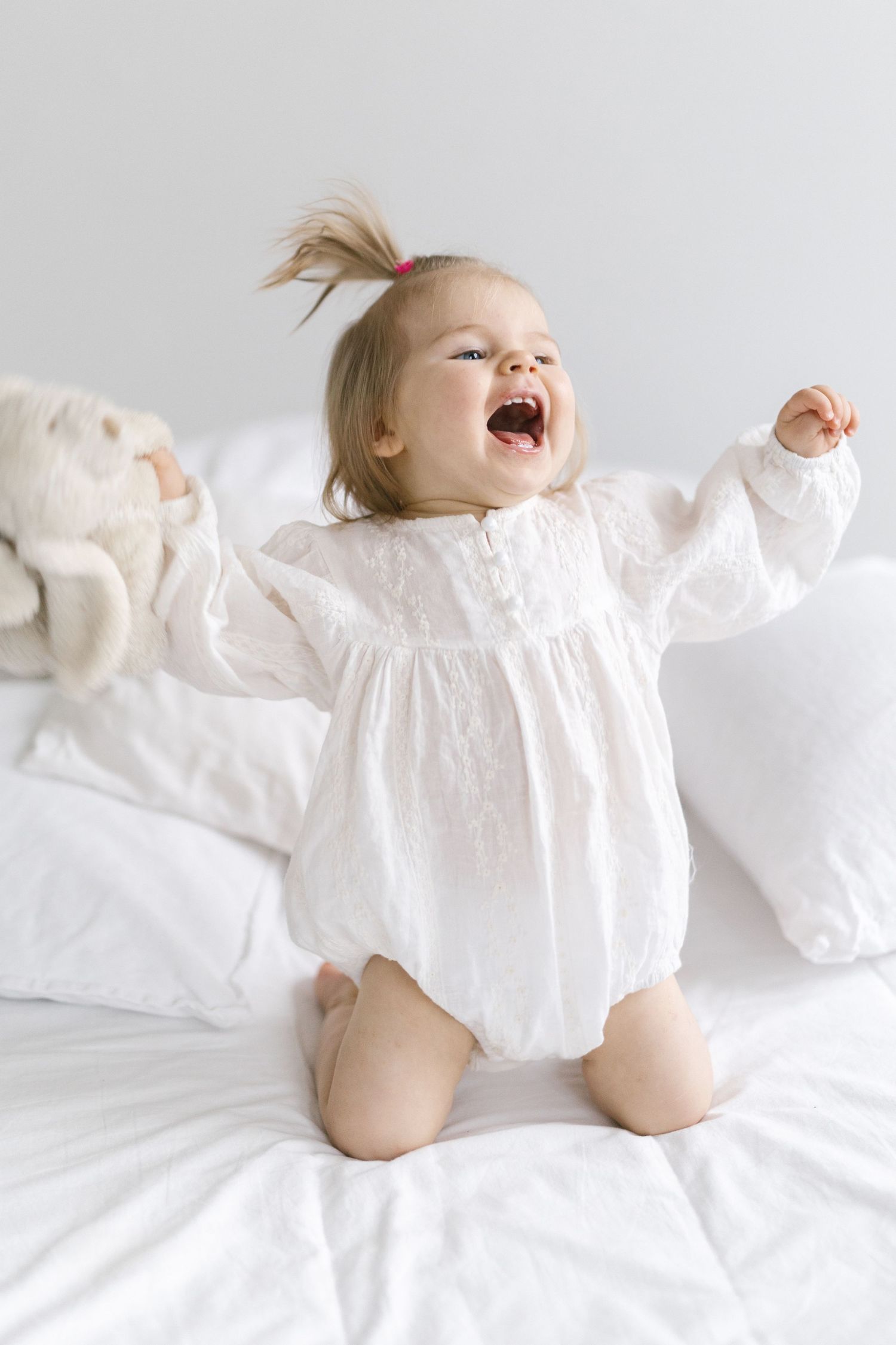 Joyful baby in white dress lying on white bedding with arms spread wide.