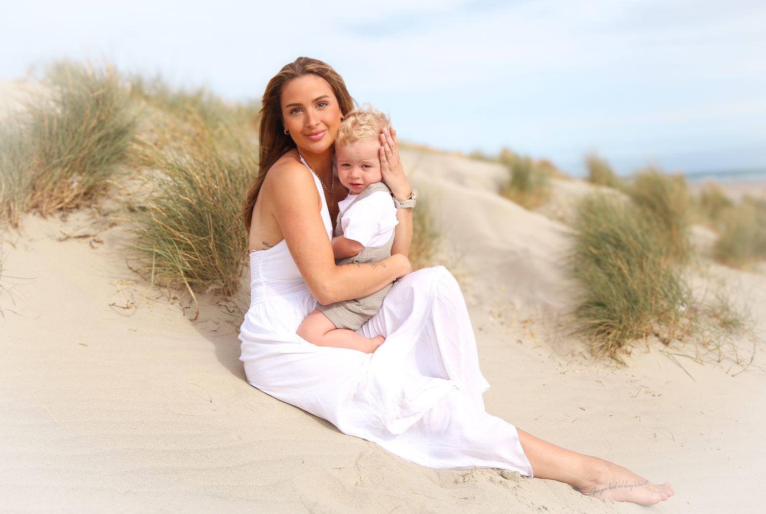 Mother and child in white outfits pose together on sandy beach dunes.